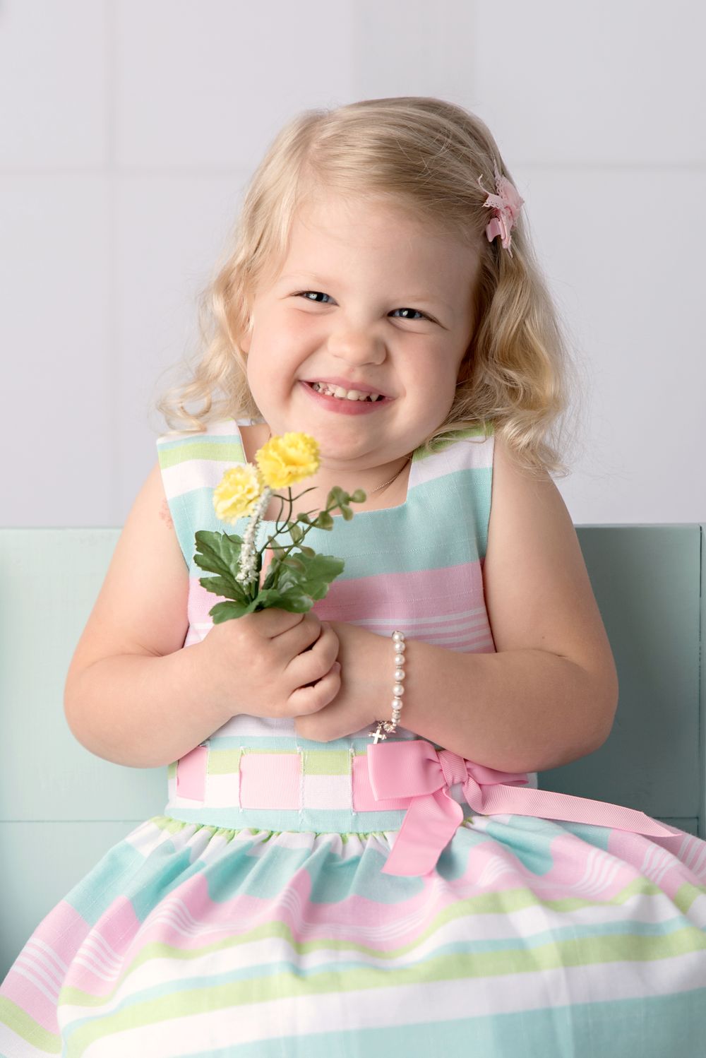 A cheerful blonde toddler girl in a pastel striped dress holds yellow roses while smiling brightly.