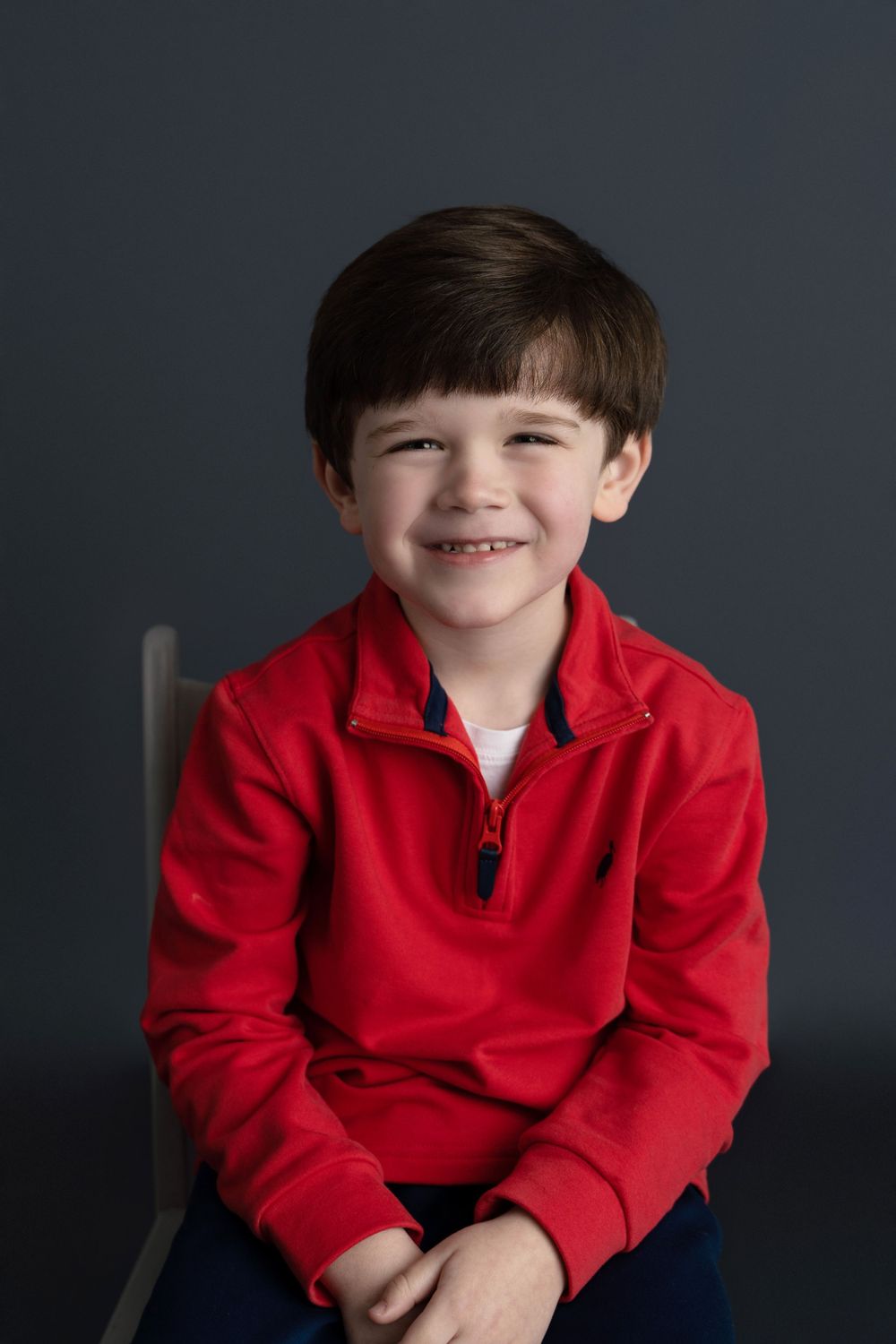 A smiling young boy in a red zip-up sweatshirt sits on a chair against a dark gray background.