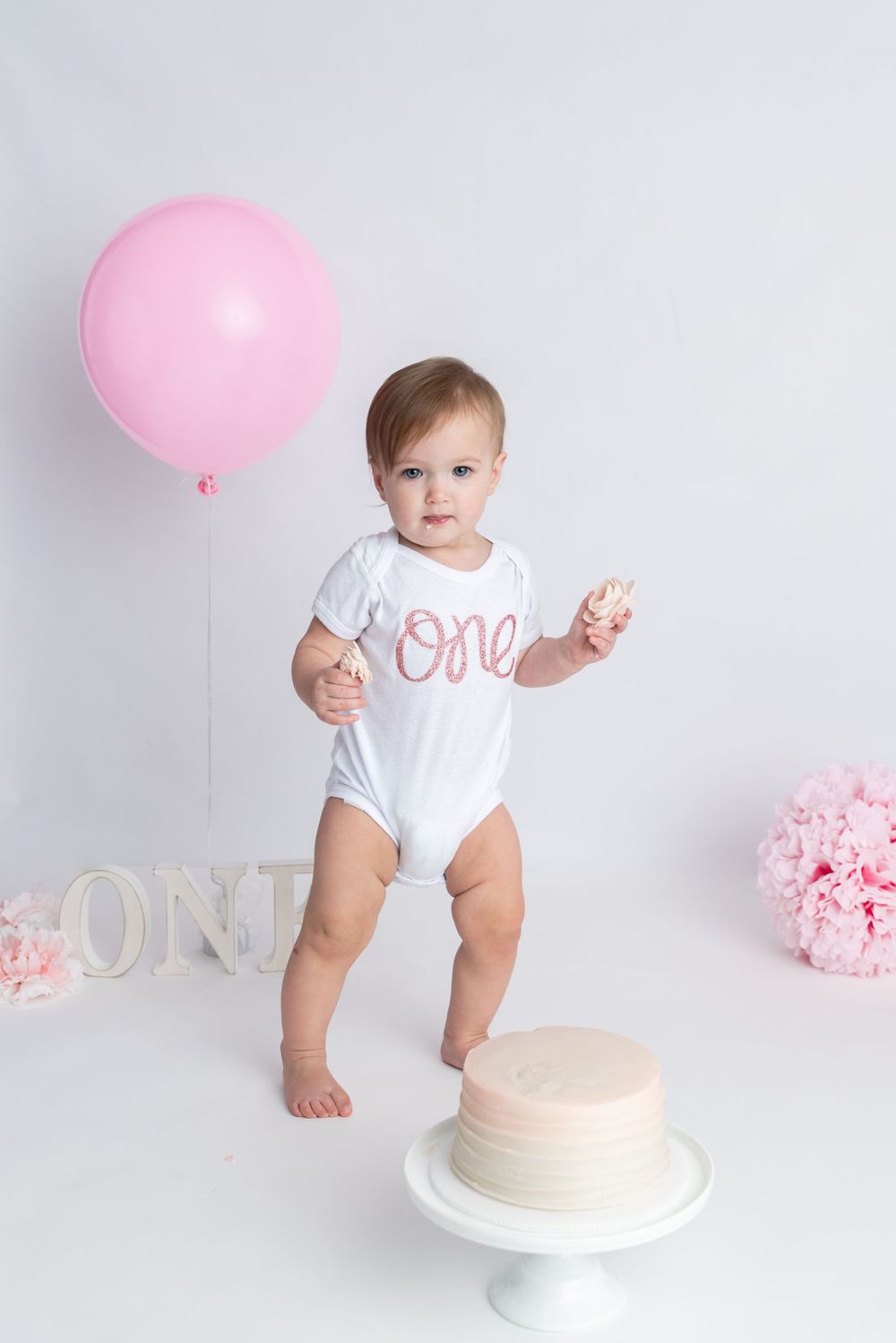 A one-year-old girl in a white onesie holds a pink balloon next to her first birthday smash cake.