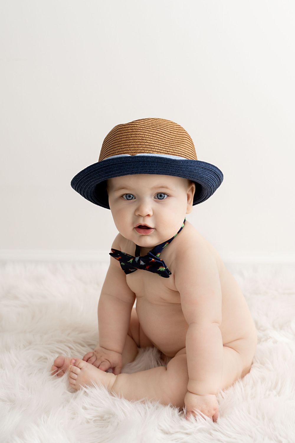 Adorable baby wearing a straw hat and bow tie sitting on a fluffy white rug in a bright studio setting.
