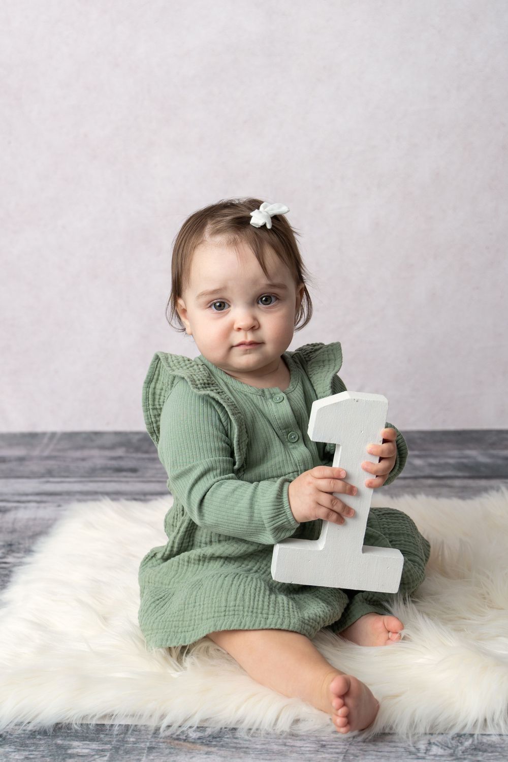 Adorable baby girl in green dress sits on fur rug holding white number one for first birthday photo session.