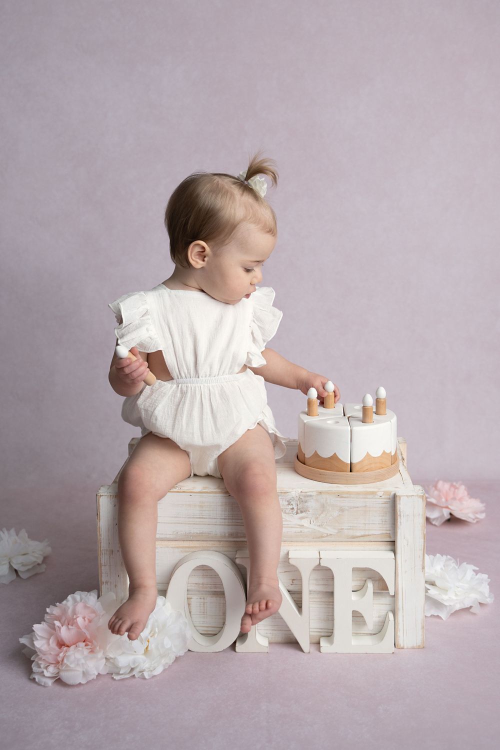 Baby girl in white romper sits on wooden crate touching first birthday cake beside ONE letters on pink background.