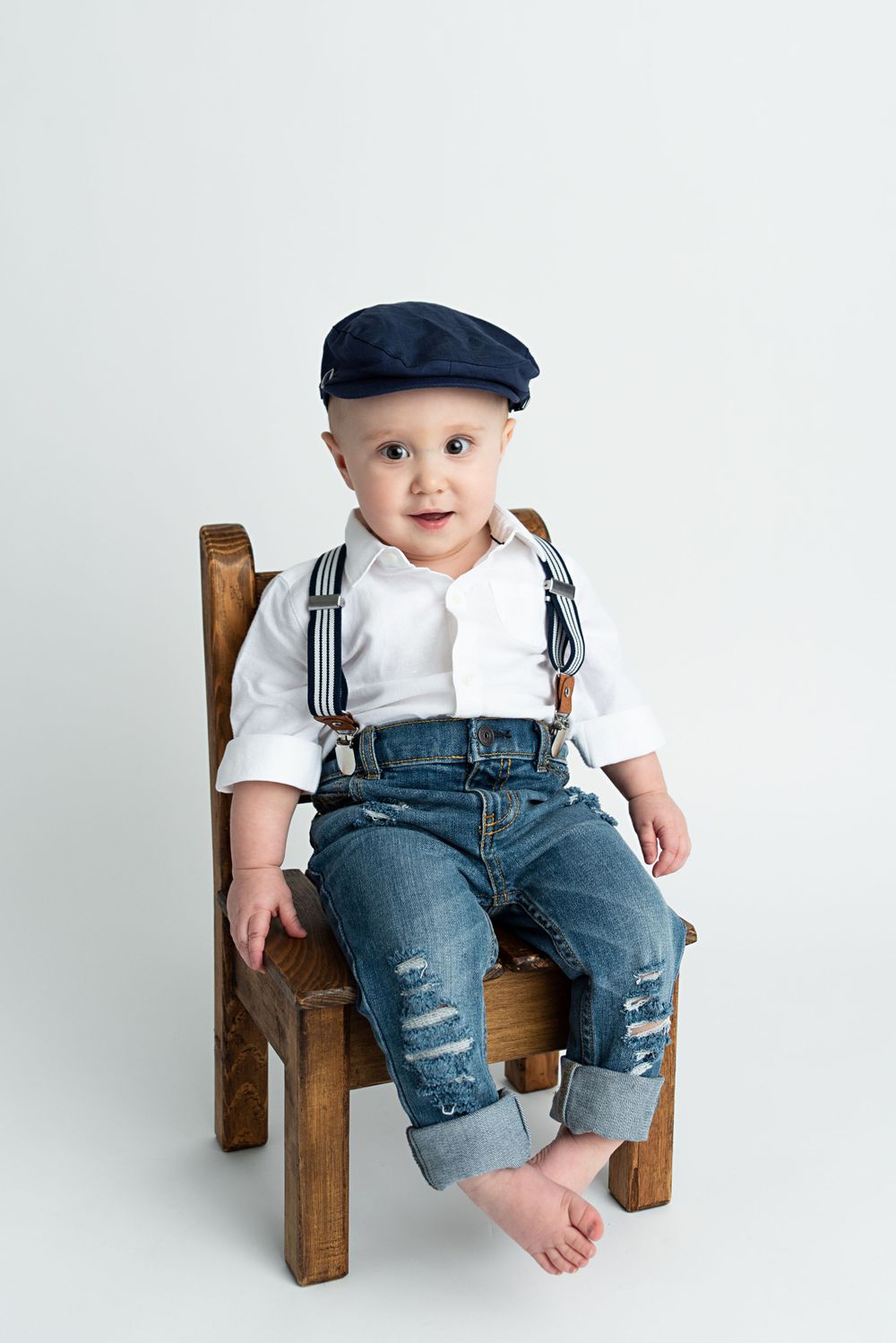 Adorable baby boy in jeans, suspenders, and newsboy cap sitting on small wooden chair.