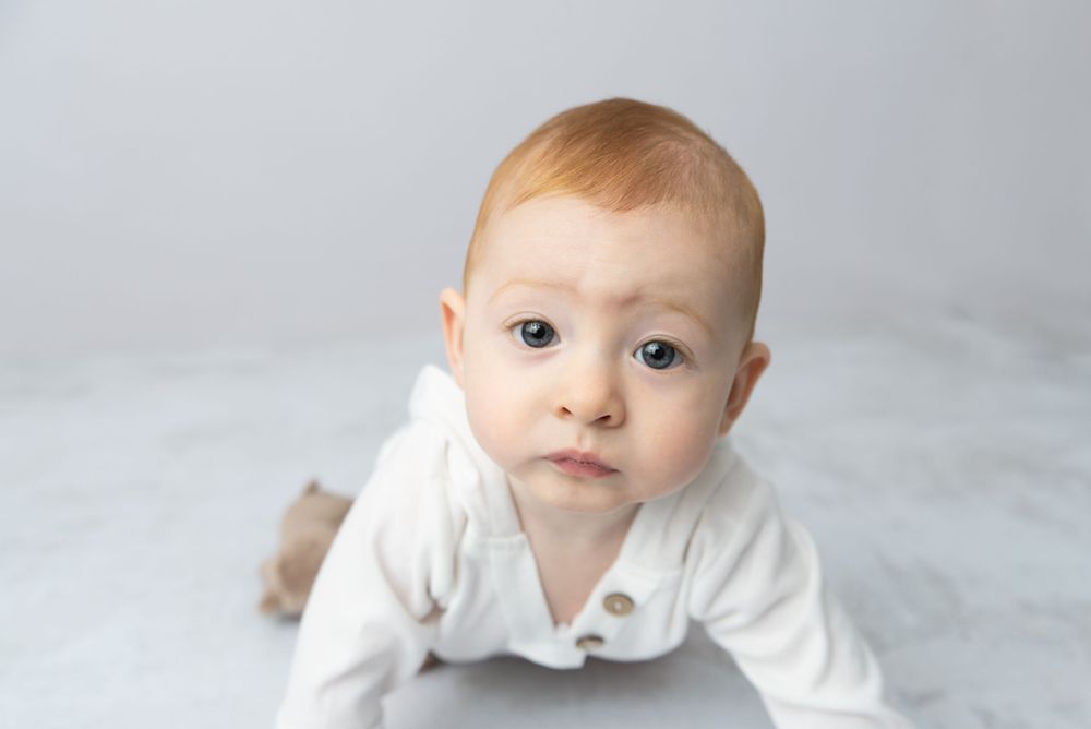 Curious red-haired baby in white onesie doing tummy time, looking up with wide eyes on a soft gray surface.