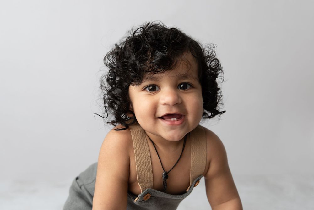 A smiling toddler with curly dark hair wearing a tan top and gray overalls poses on a white background.