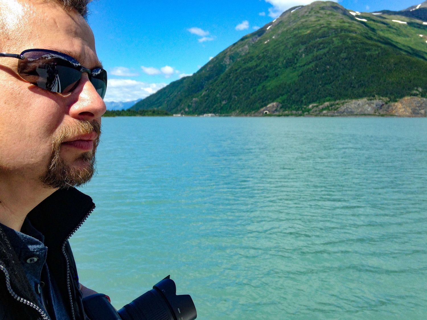 Person in sunglasses beside water with a mountain in background.