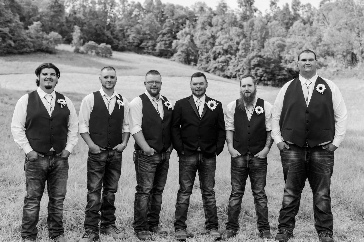 Wedding party in casual western attire posed in black and white on grassy hillside.
