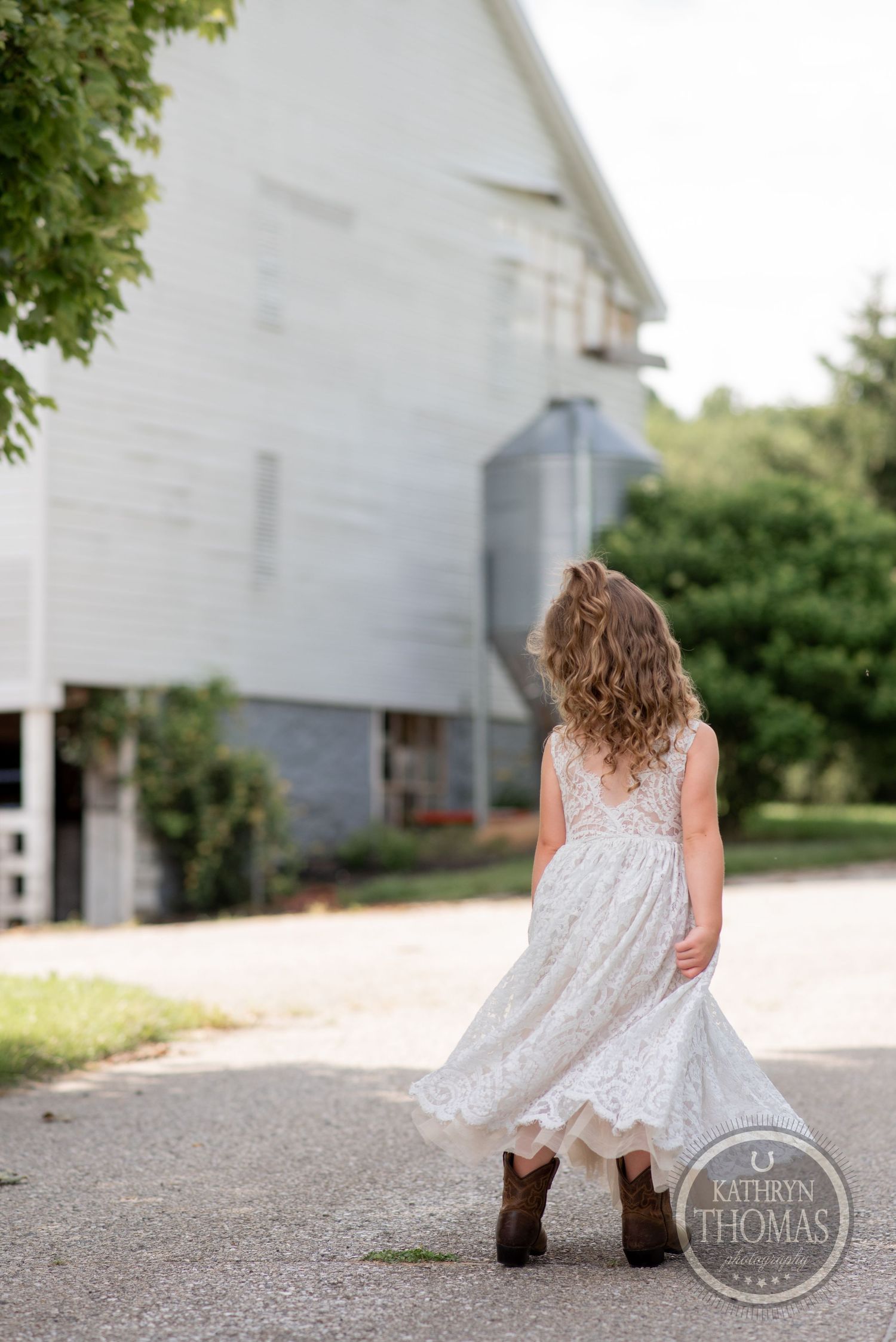 Young girl in white lace dress and cowboy boots twirls near a rustic white barn.