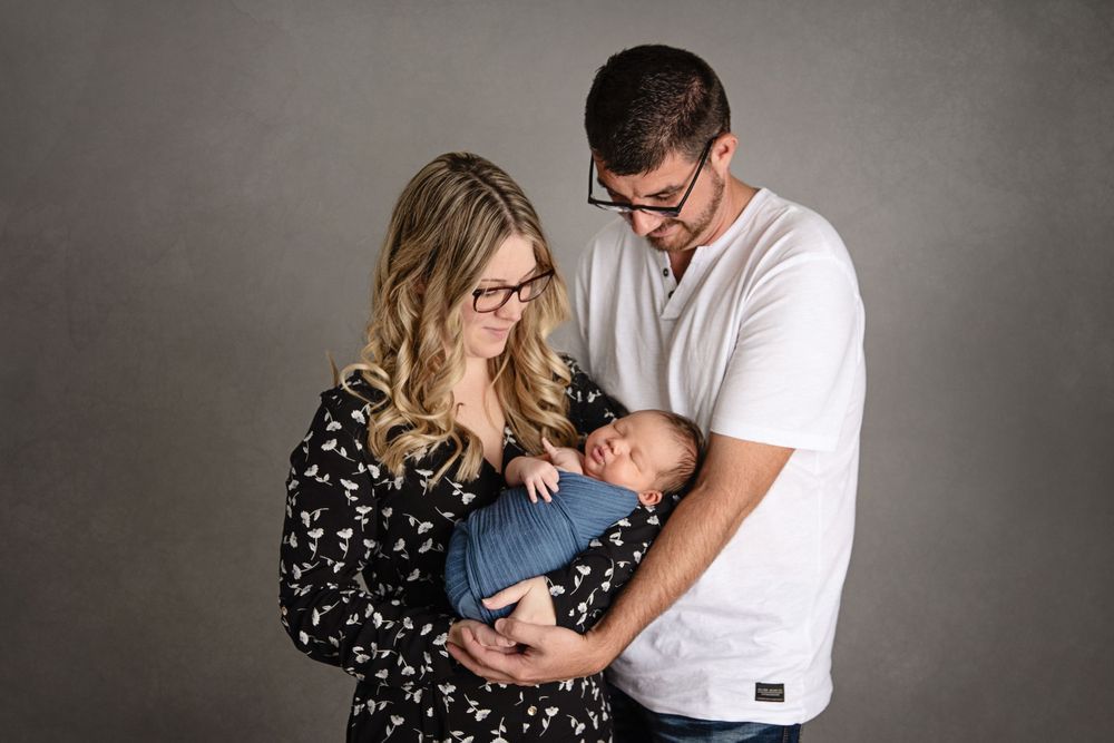 Happy couple holding their newborn baby wrapped in blue blanket against grey background.