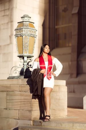 Graduate in white dress and red stole poses by vintage lamp post on campus steps.