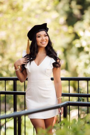 Graduate in white dress and cap poses outdoors against natural background.