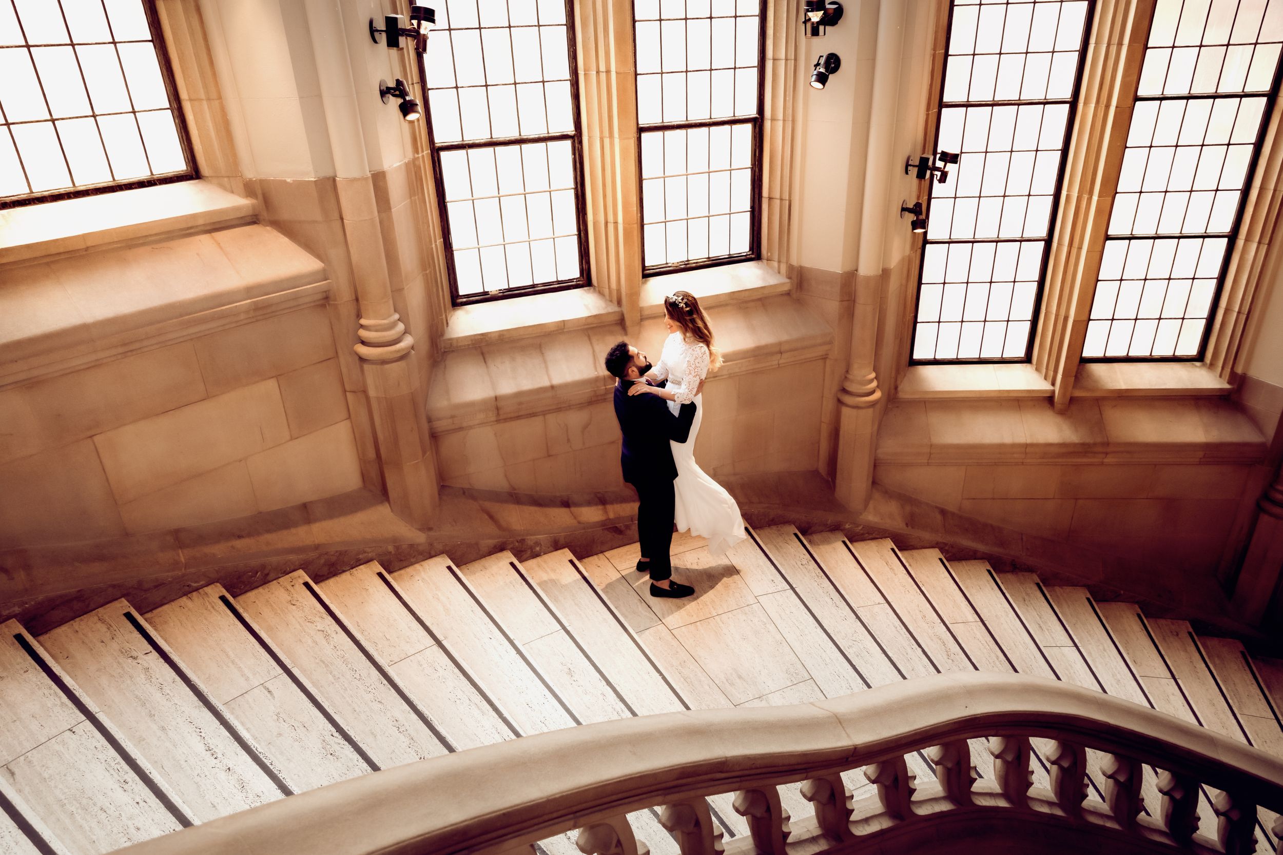 Couple embracing on historic stone staircase near ornate windows.