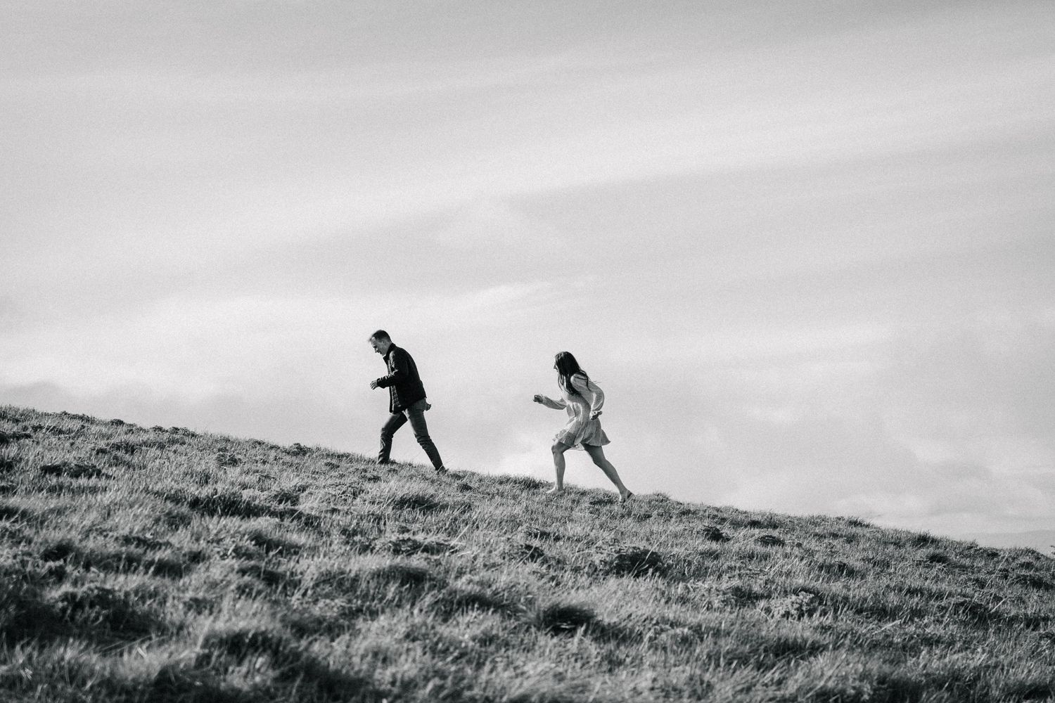 Silhouetted figures running playfully up a grassy hillside against cloudy sky.