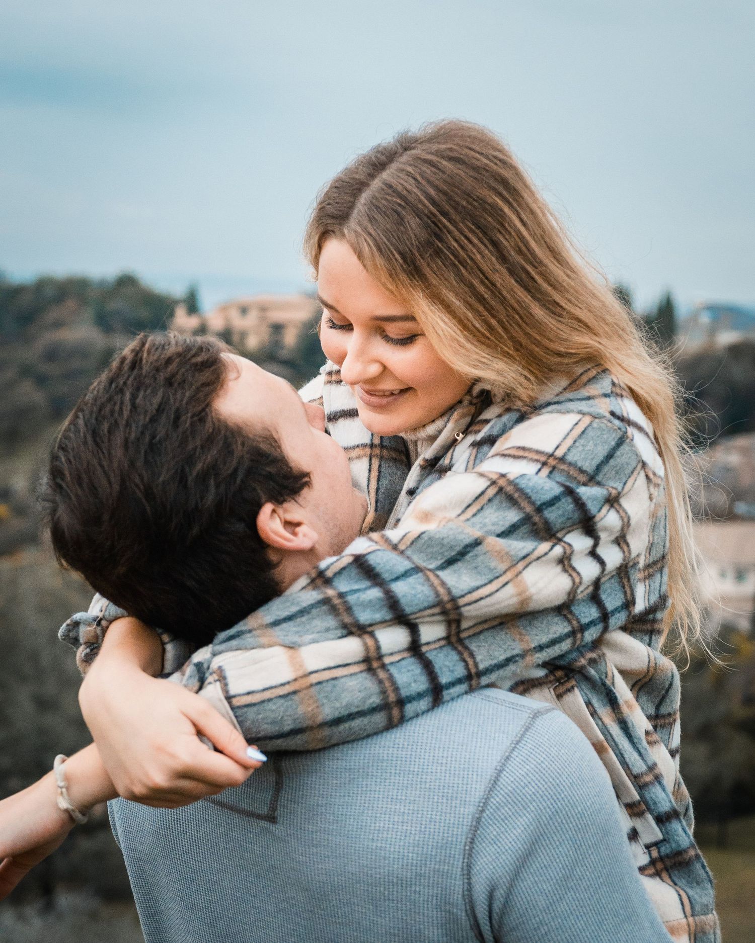 A romantic couple embraces while wearing casual clothing outdoors with a blurred landscape background.