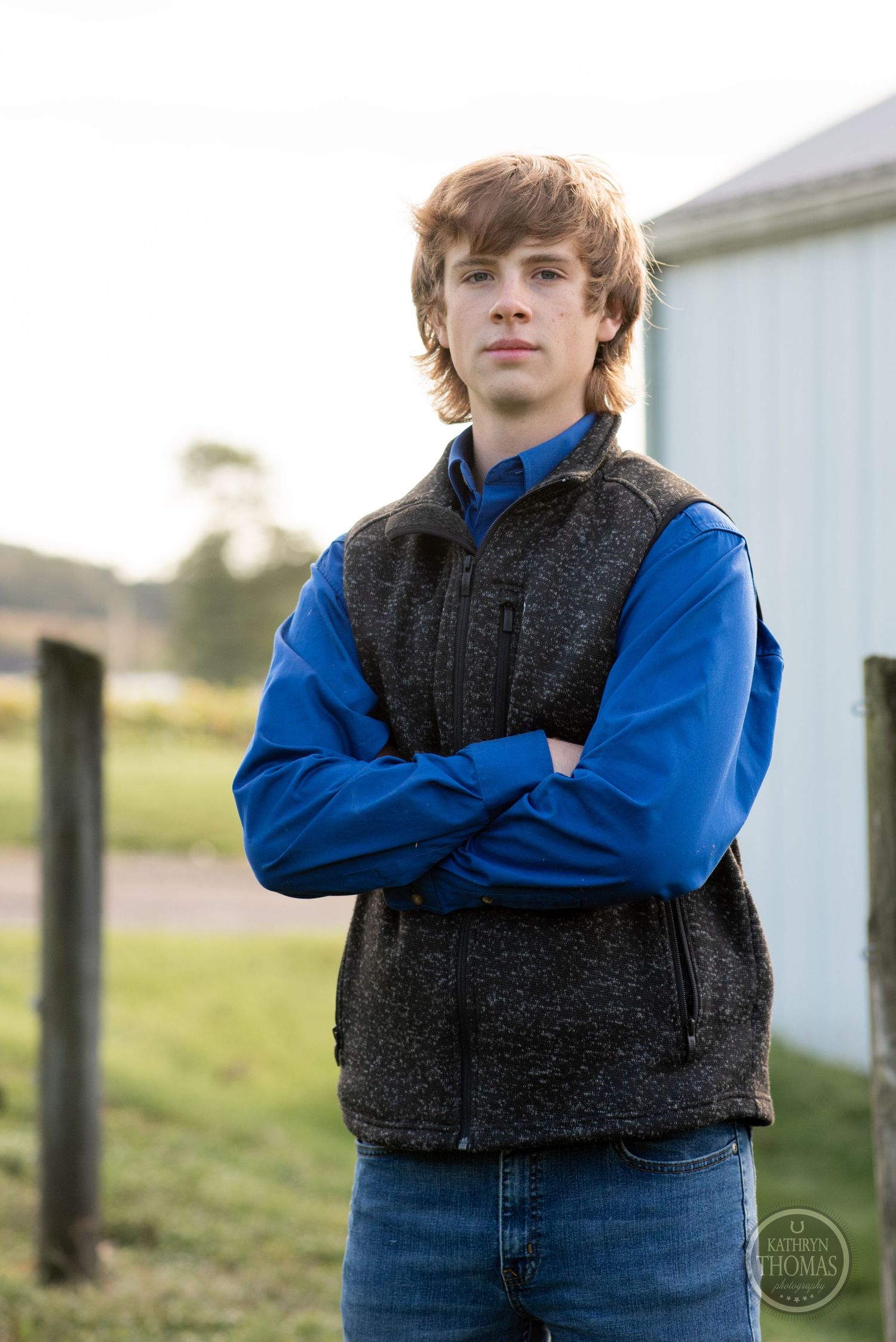 Figure in fleece vest and blue sleeves stands confidently with arms crossed by barn.