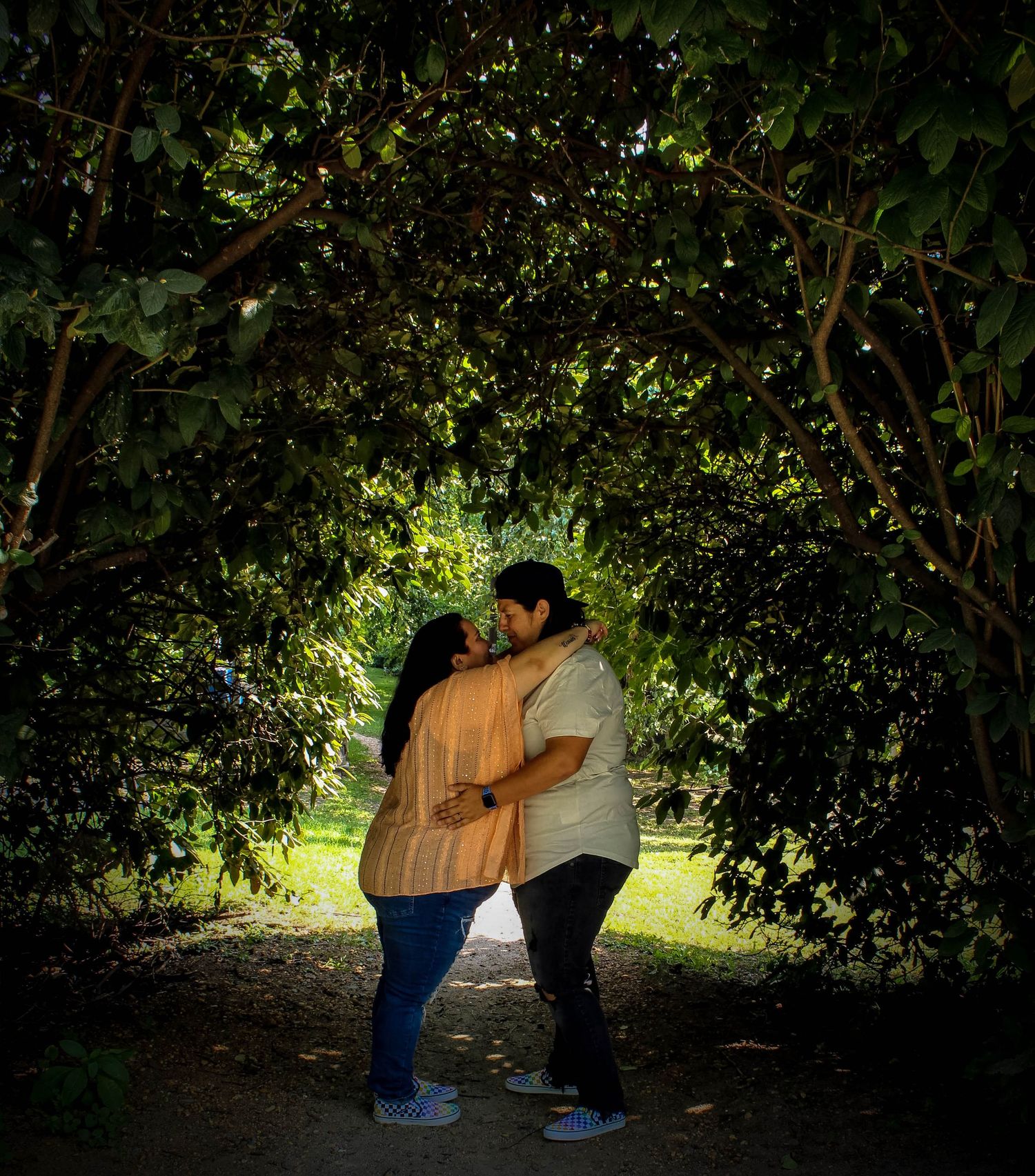 Couple embracing under tree canopy in dramatic backlit garden setting.