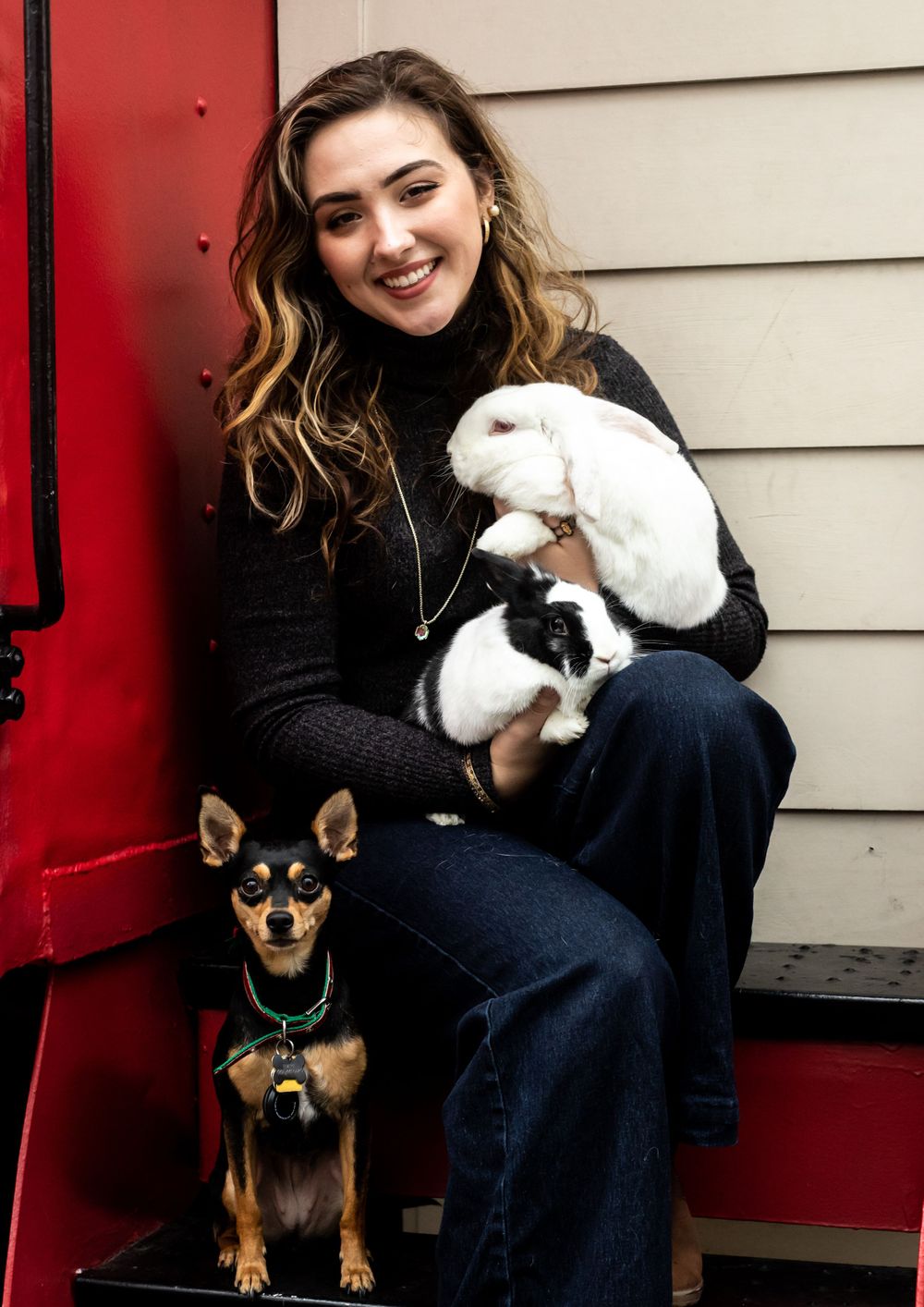 Young woman smiling while holding white rabbits with a small Chihuahua dog sitting beside her on steps.