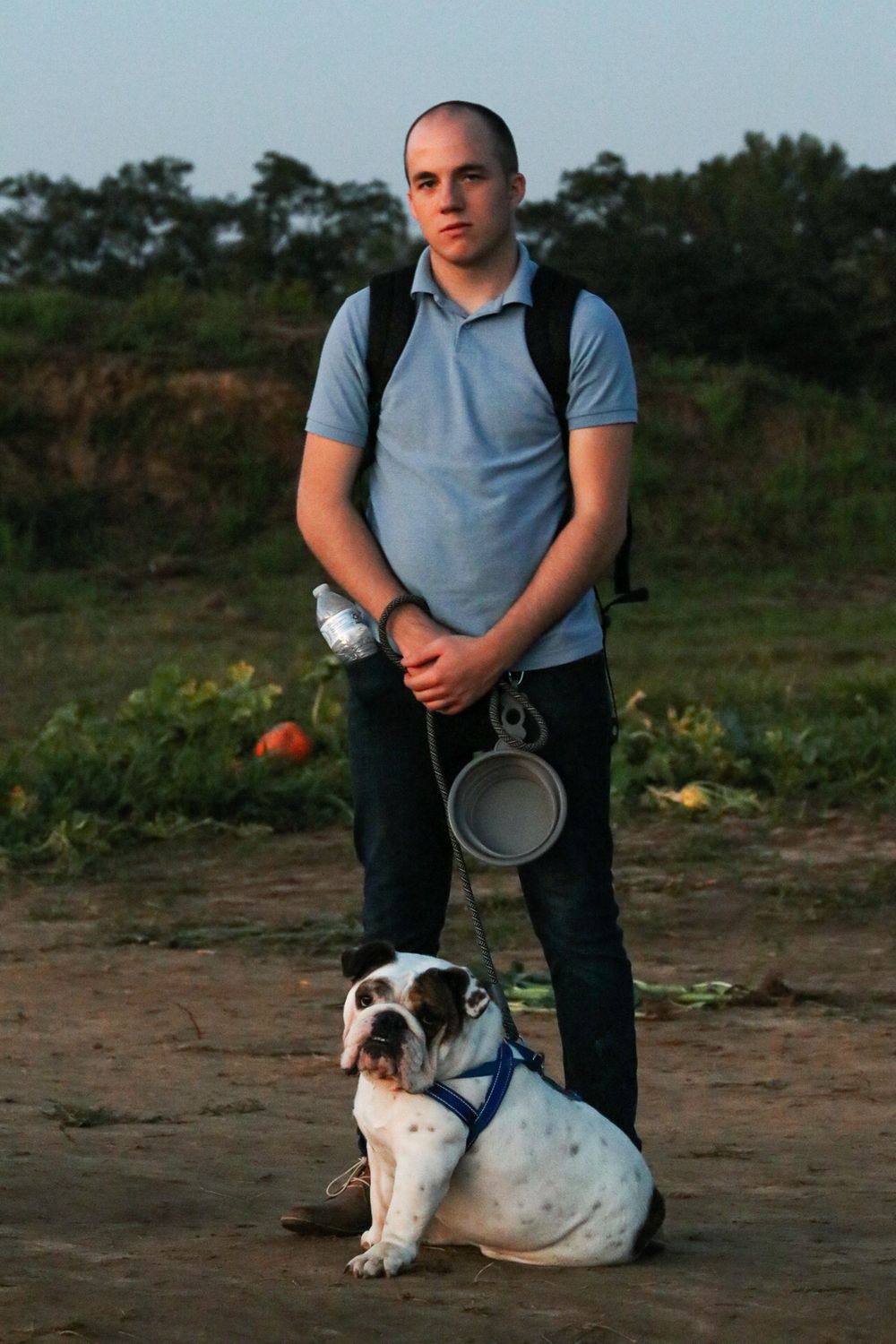 Young man with backpack standing outdoors holding leash of English Bulldog sitting on dirt path in field.