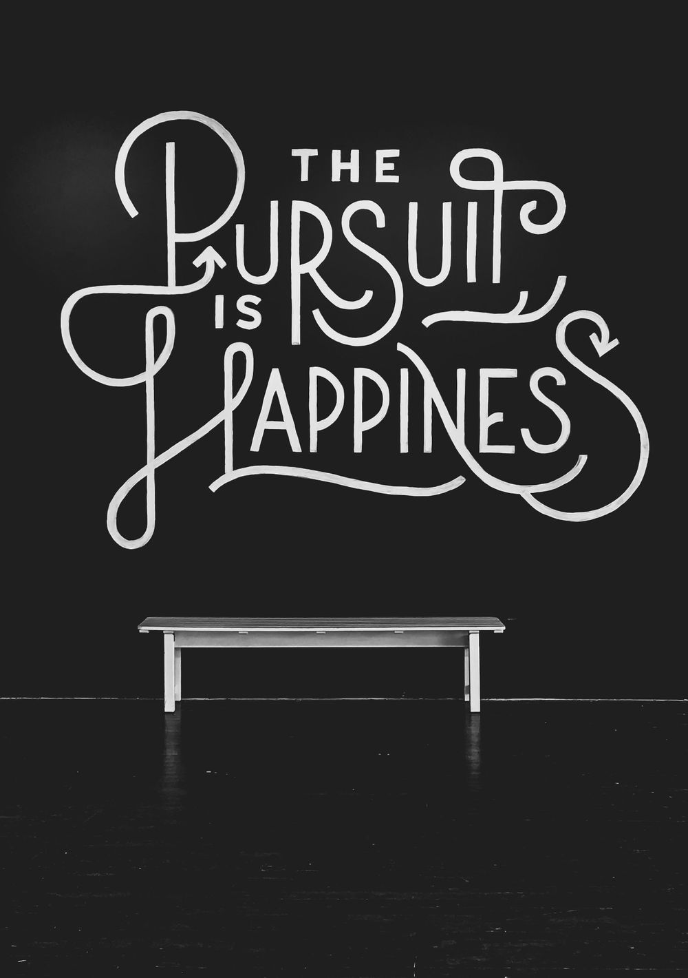 Black wall with white typographic art reading The Pursuit is Happiness above a bench.
