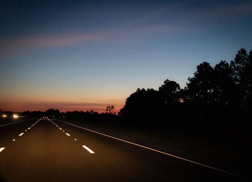 Empty highway at dusk with colorful sunset sky and silhouetted trees.