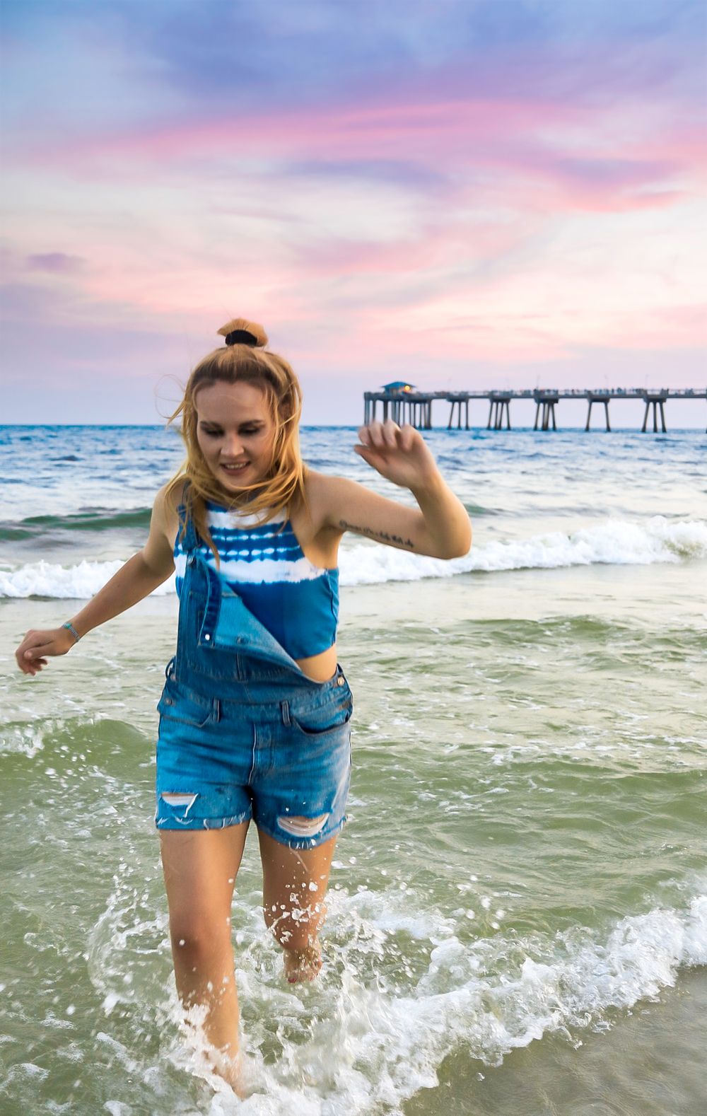 Young blonde woman in denim overalls joyfully splashing in ocean waves at sunset beach.