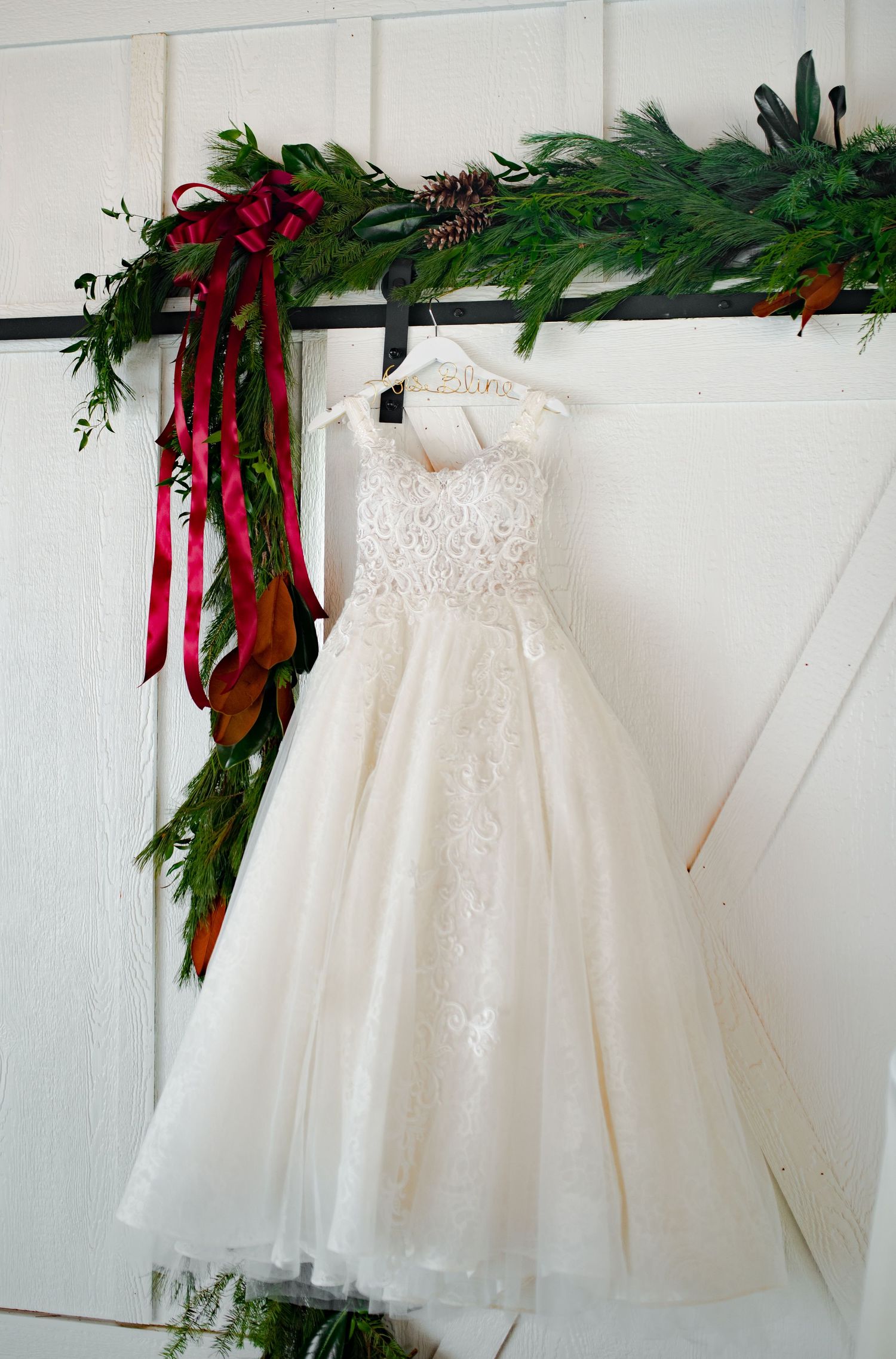 Delicate white christening gown with lace details hangs on display with festive red ribbons and greenery above.