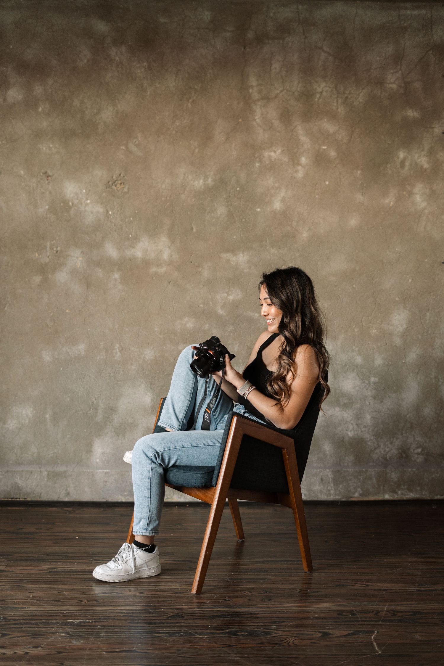 A photographer sitting in a wooden chair against a textured wall with a professional camera.