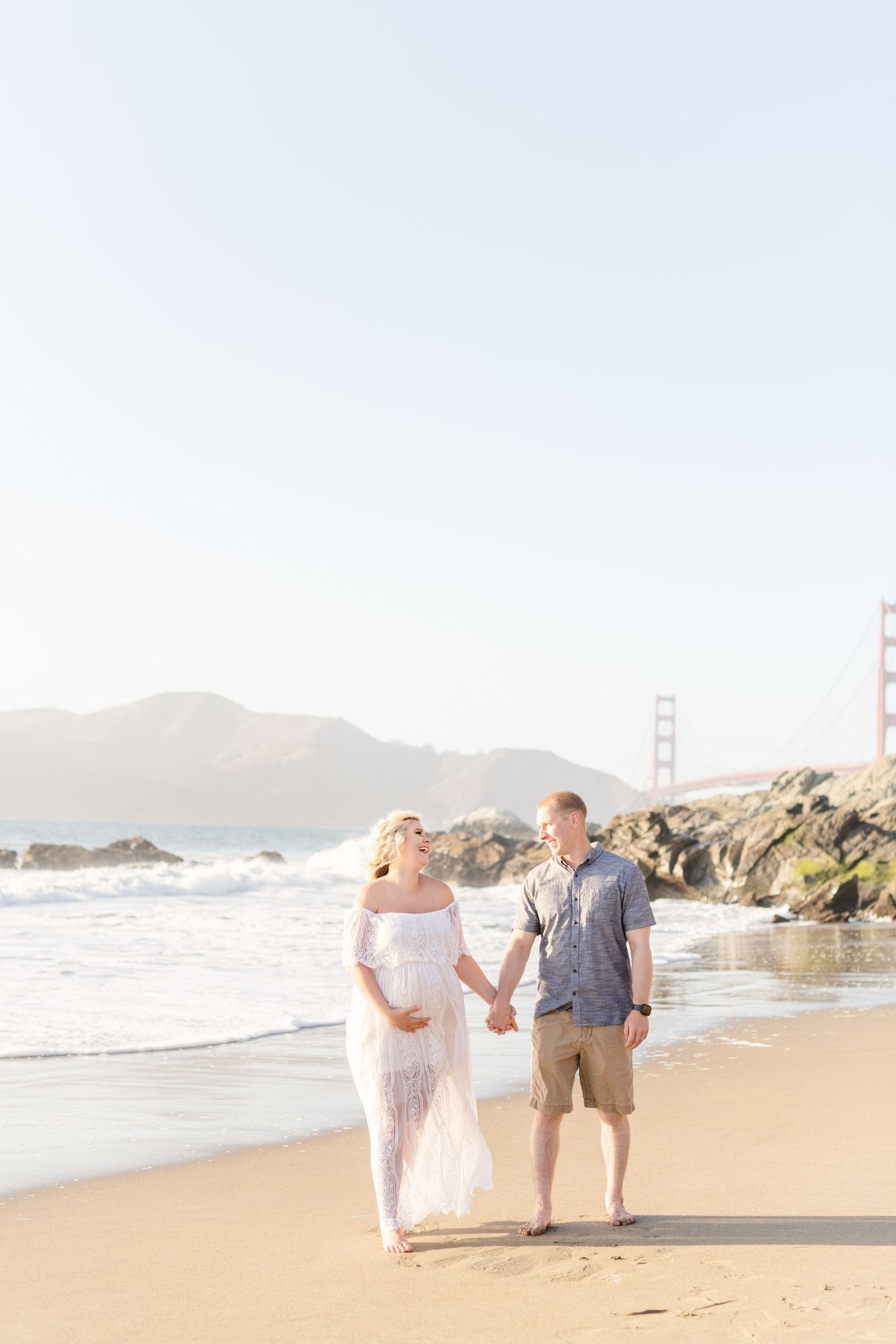 Couple walking barefoot on beach with Golden Gate Bridge visible in soft lighting.