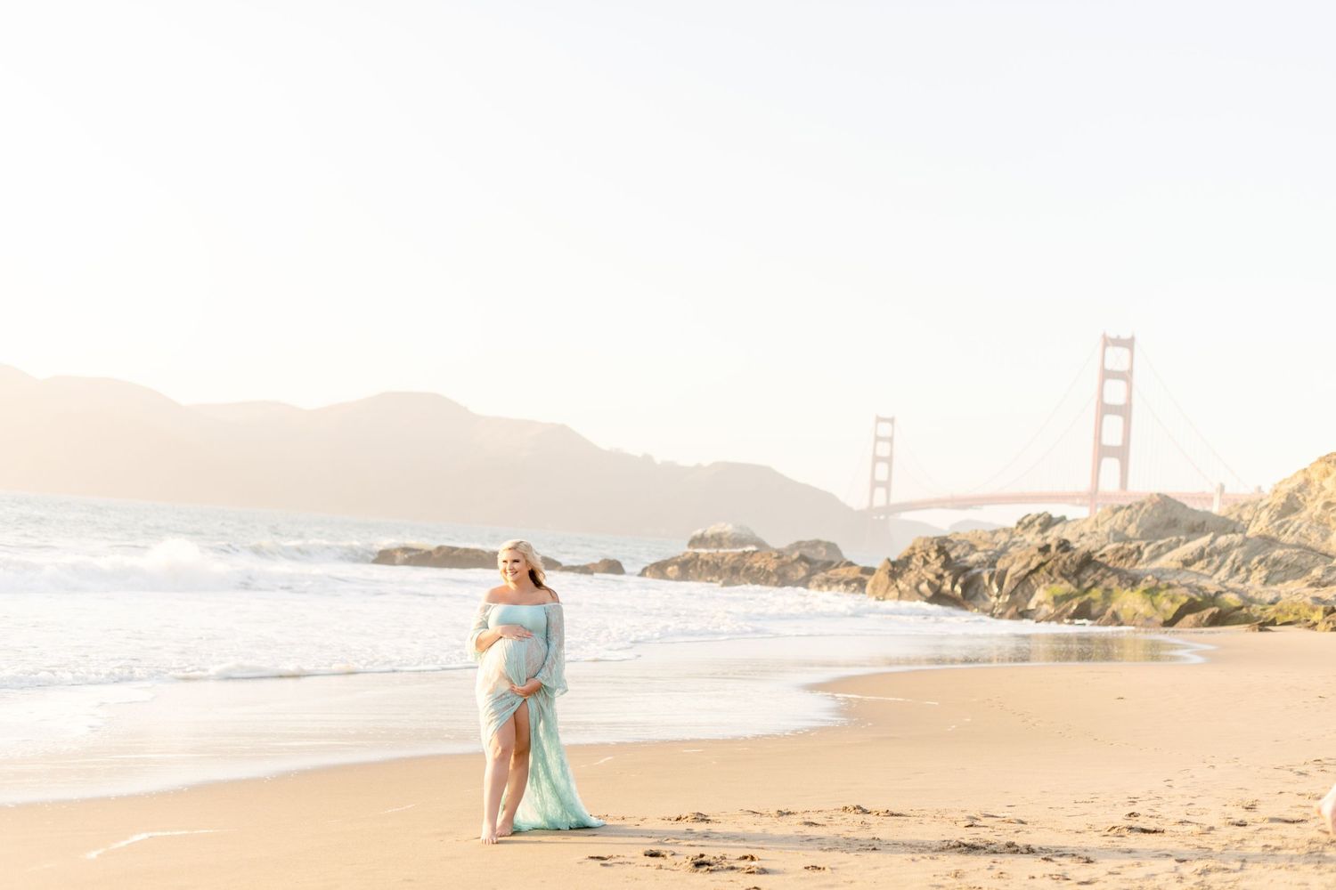 Figure in light blue dress poses on beach with Golden Gate Bridge in background.