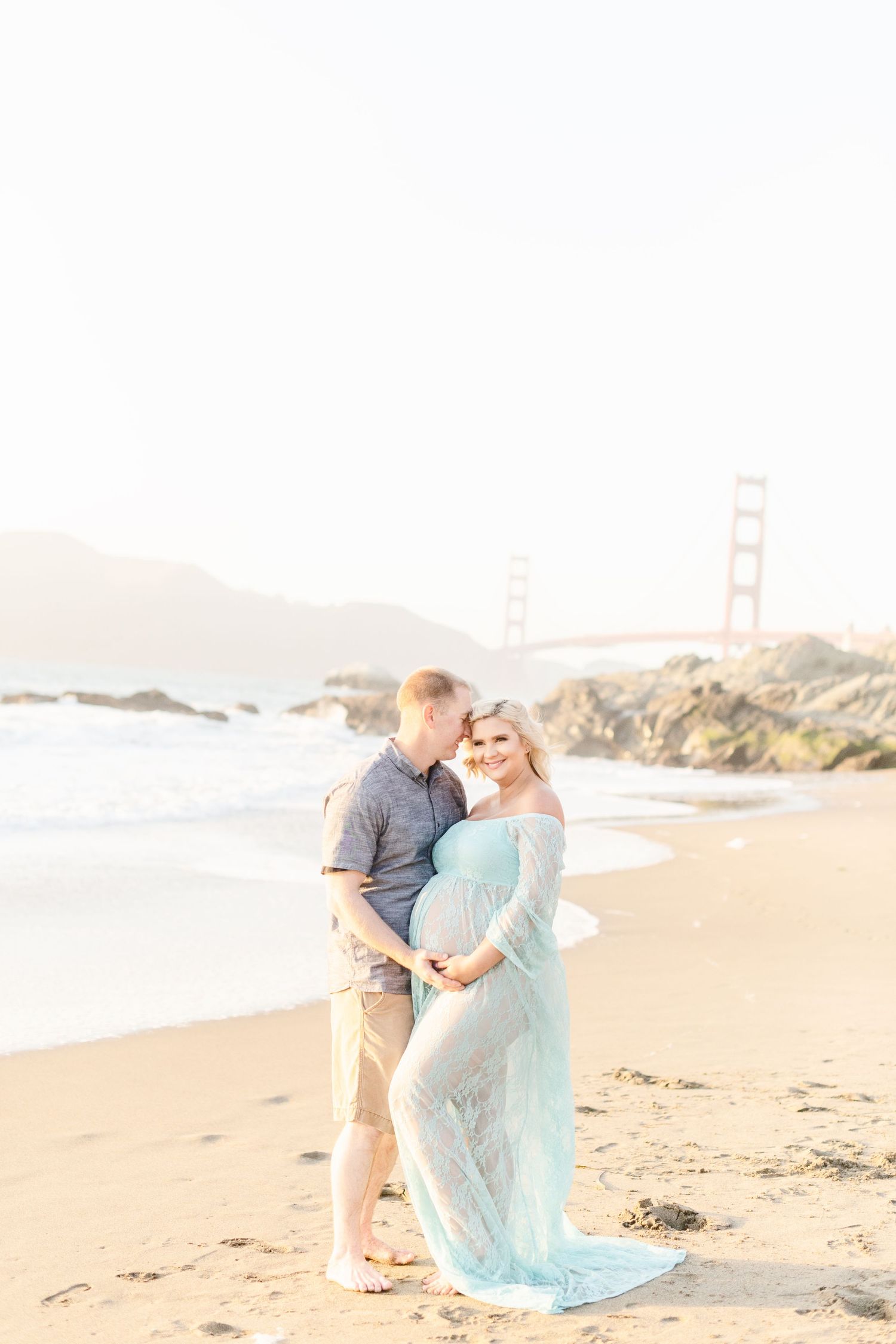Maternity photo session on sandy beach with iconic bridge visible in distance.