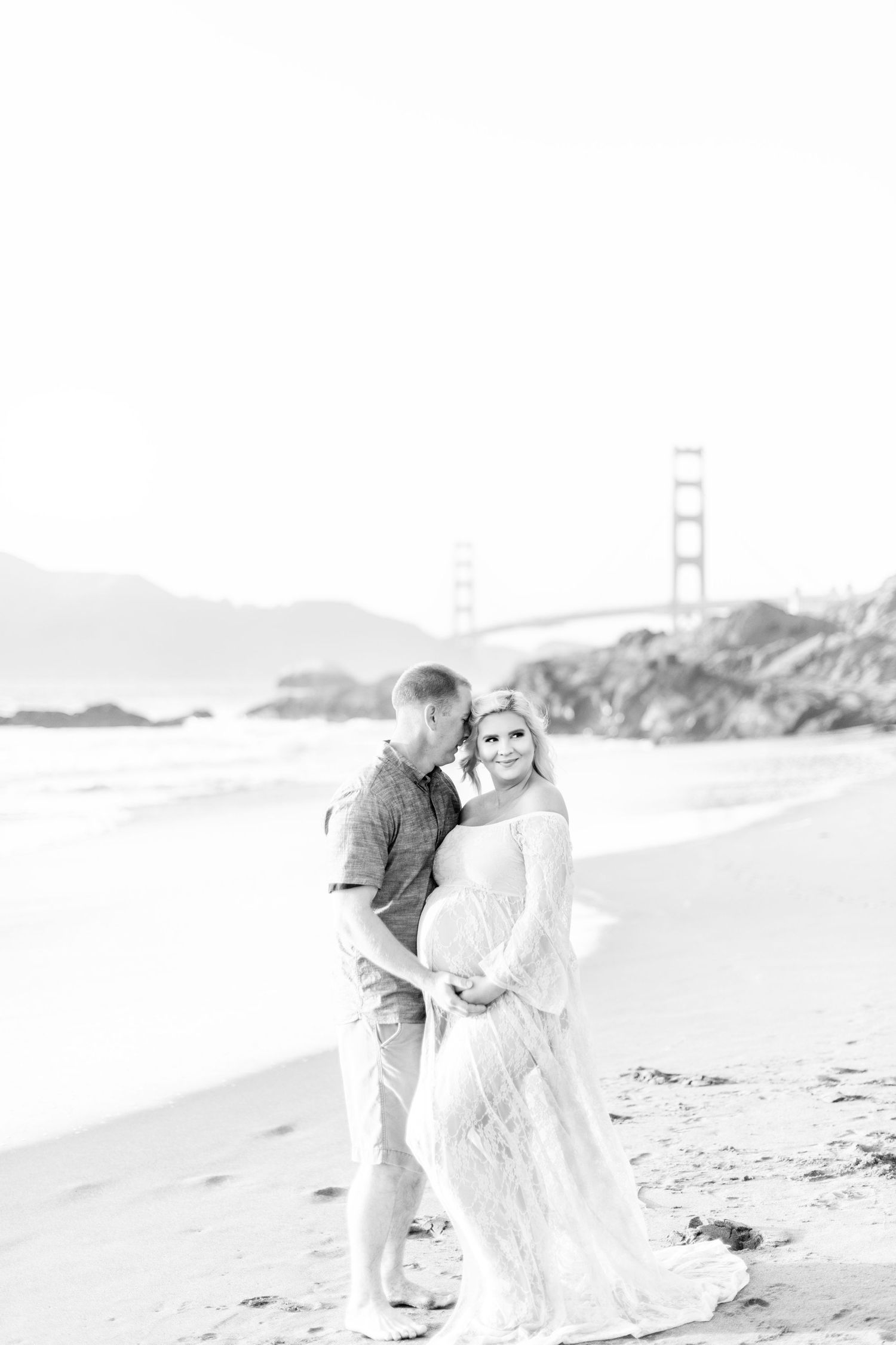 Romantic couple embraces on beach with Golden Gate Bridge visible in black and white photo.