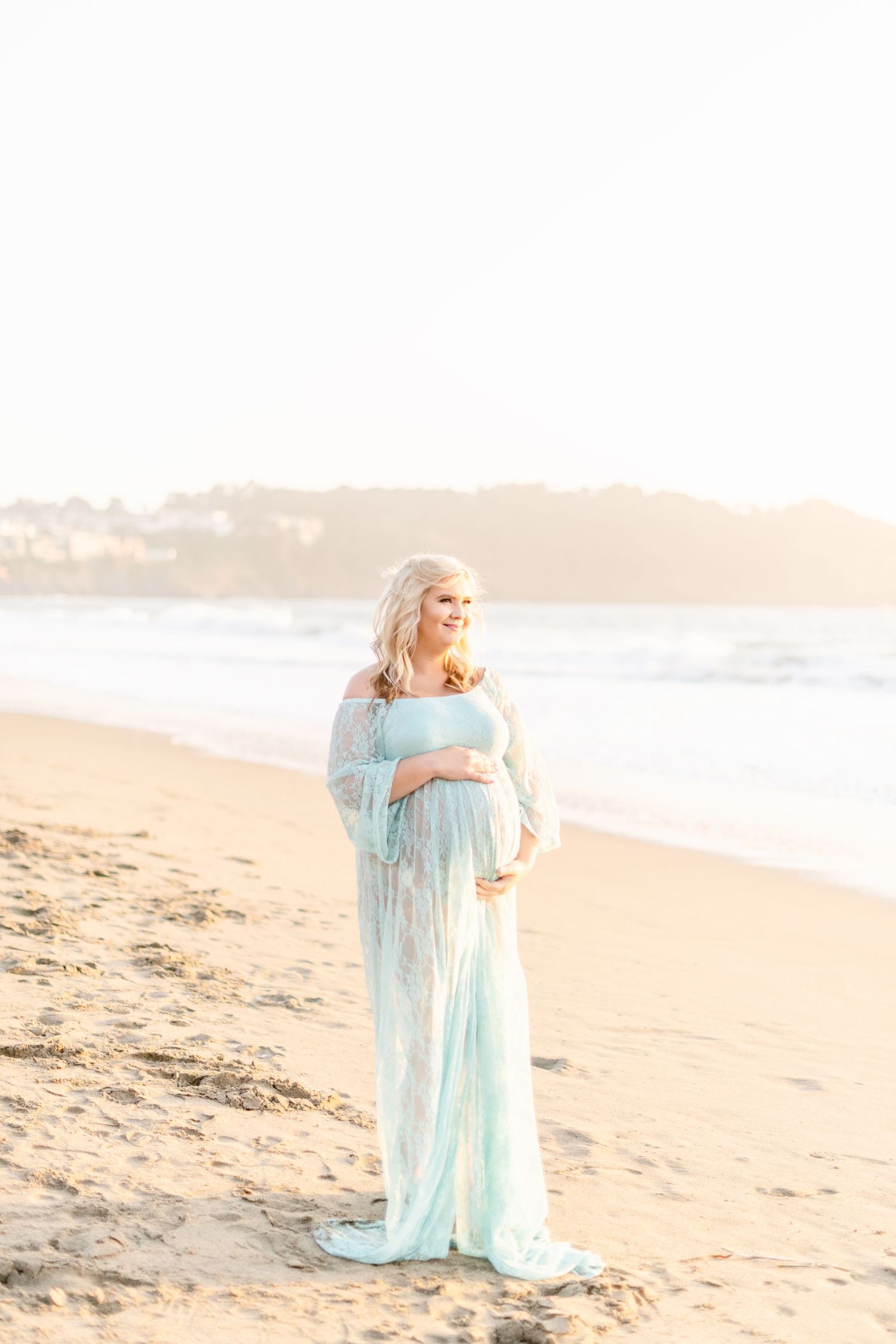 Woman in ethereal blue gown stands on beach during golden hour.