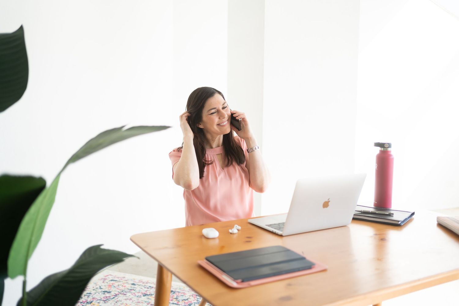 Woman smiling while working on laptop at desk with plant and water bottle.