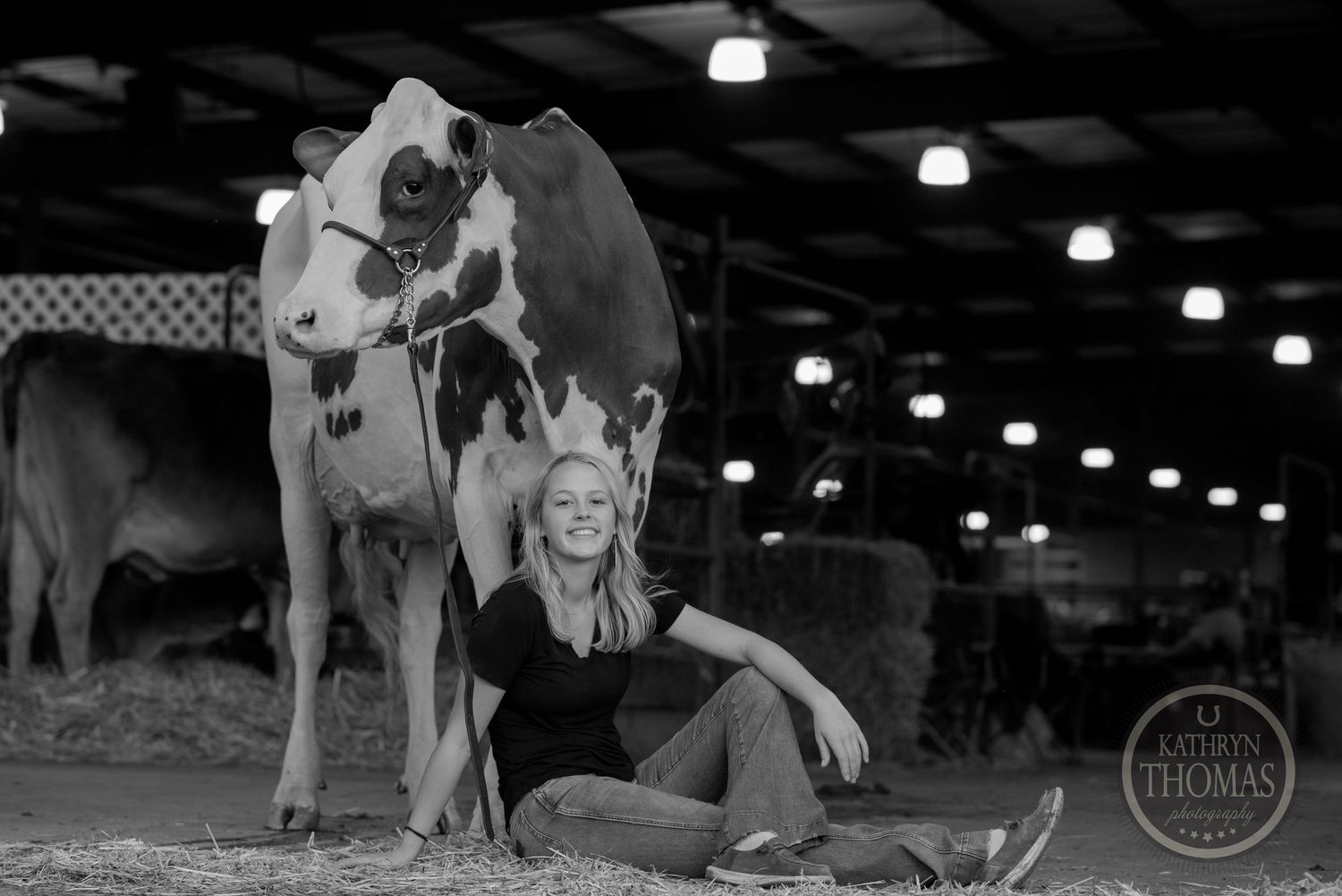 A black and white photograph of someone posing with a spotted dairy cow in a barn setting.
