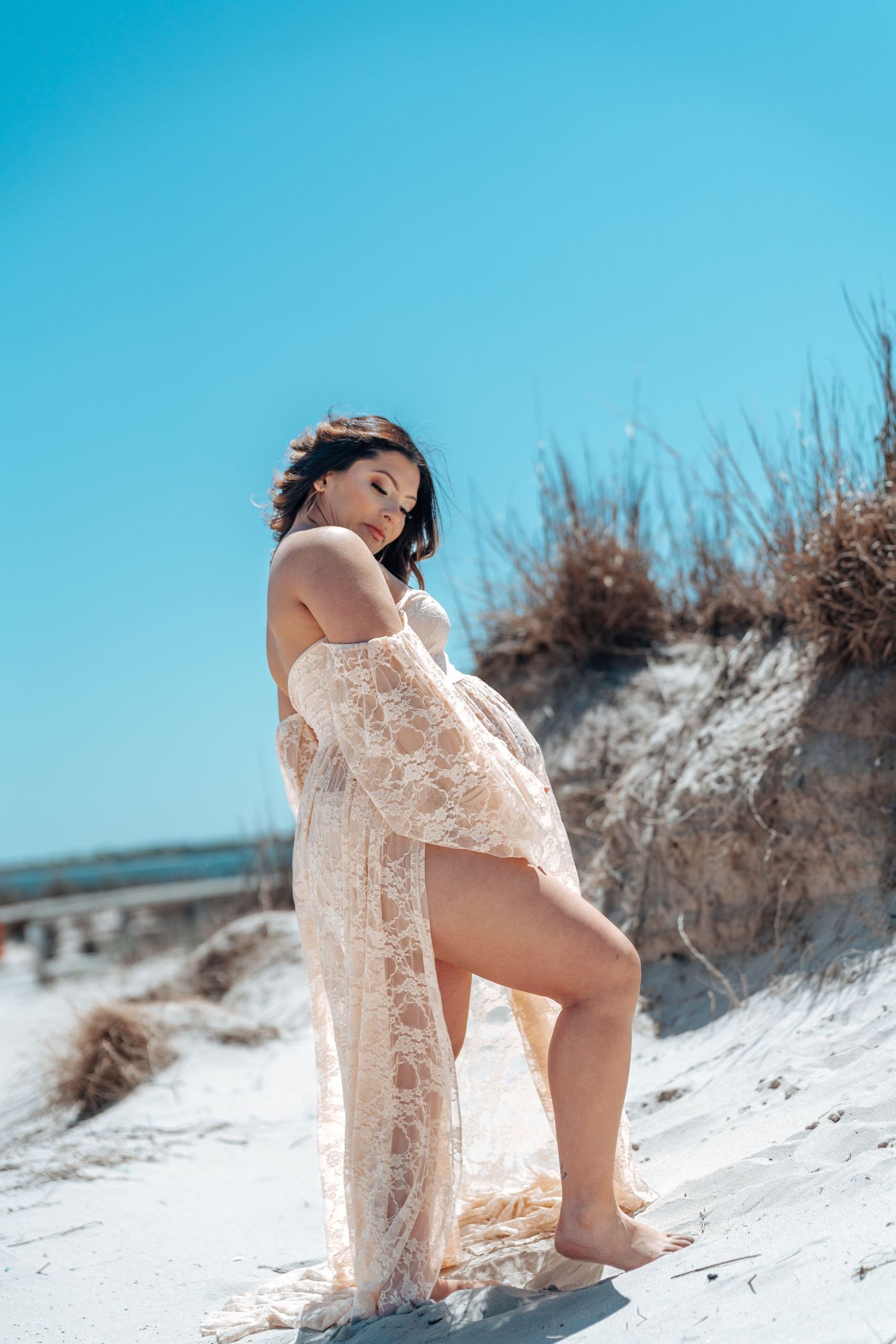 Expectant mother in a lace dress poses gracefully on white sand dunes against vibrant blue sky.