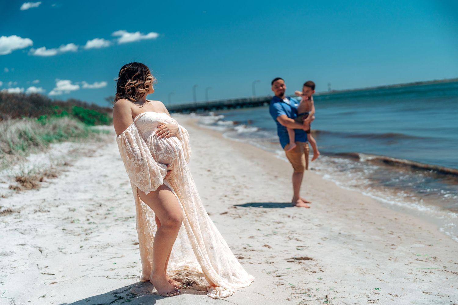 Maternity photoshoot on beach with family silhouettes in background.
