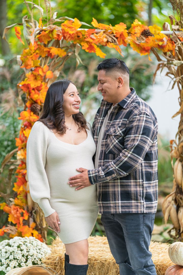 A maternity photo session with a rustic fall-themed backdrop decorated with orange leaves and hay bales.