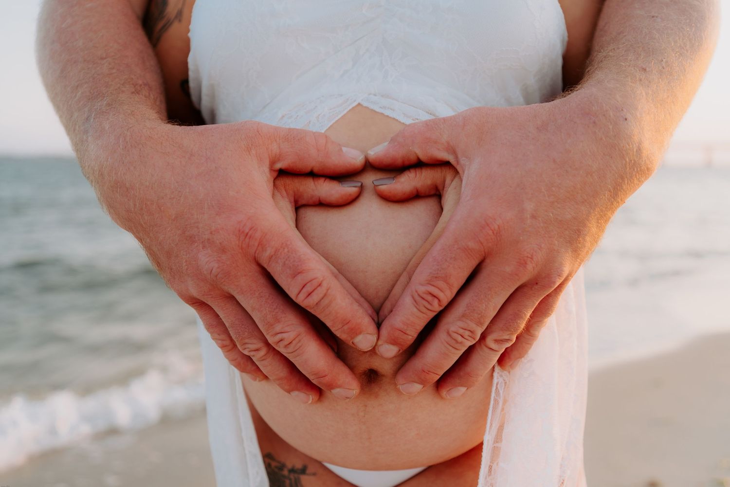 Hands form heart shape over pregnant belly on beach at sunset.