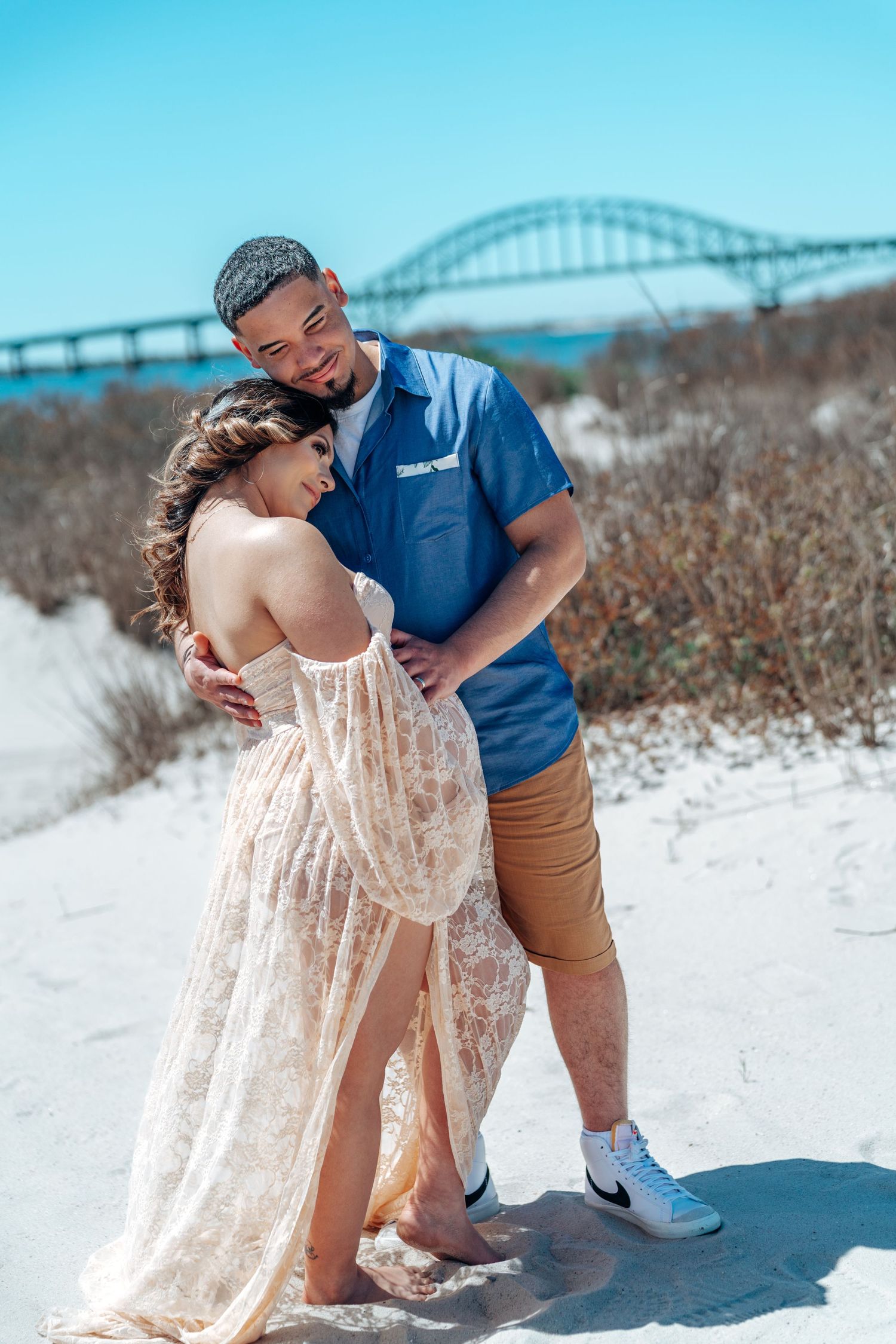 Couple embraces on sandy beach with bridge visible in background during sunny day.