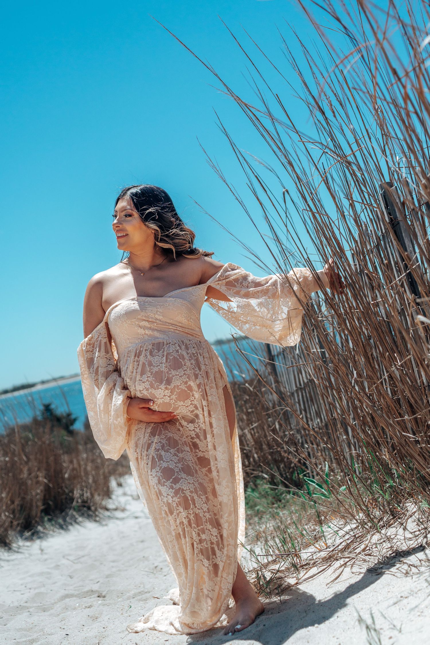 Expecting mother in flowing lace dress poses by beach grass under bright blue sky.