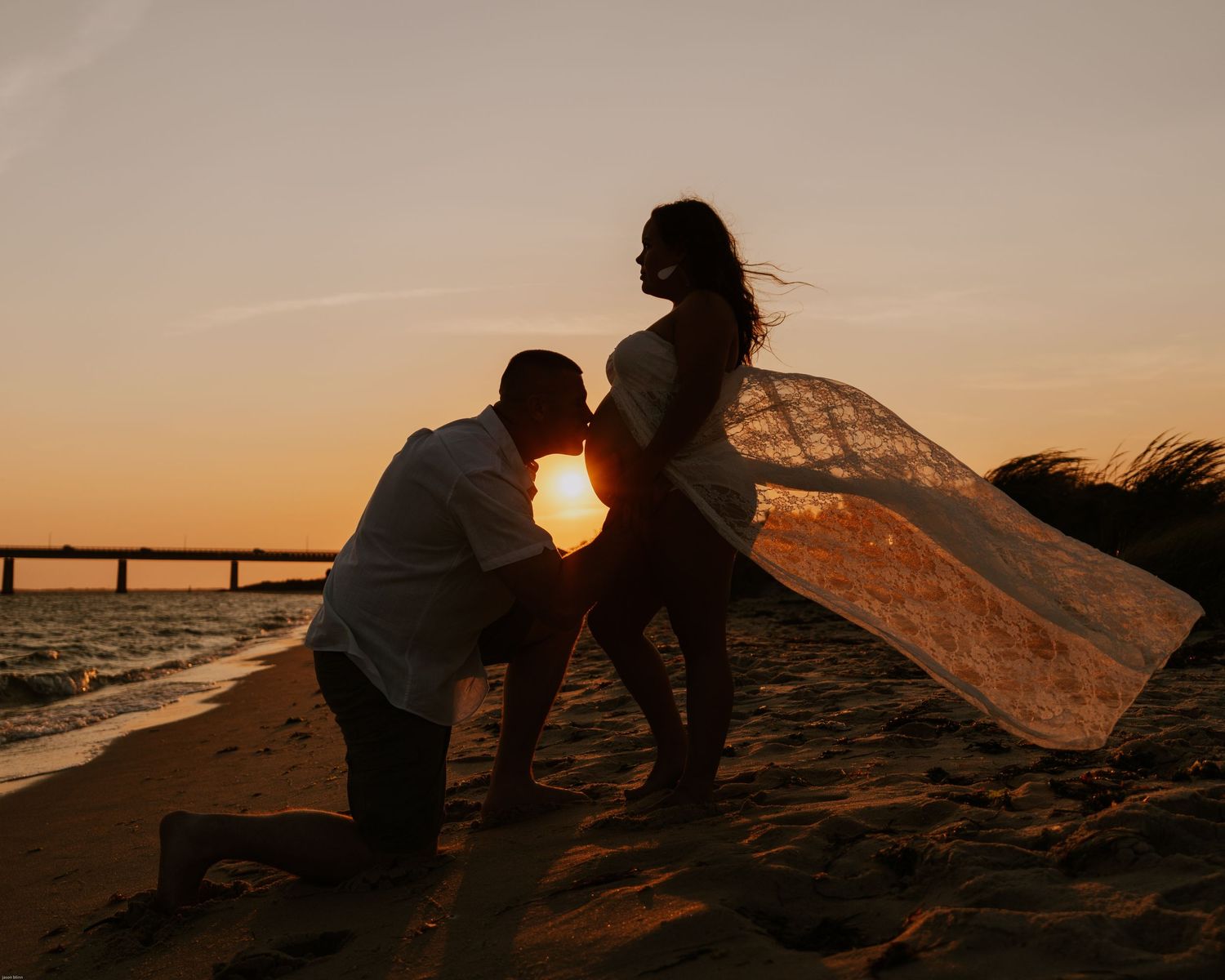 Silhouette of couple during sunset maternity shoot on beach with bridge in background.