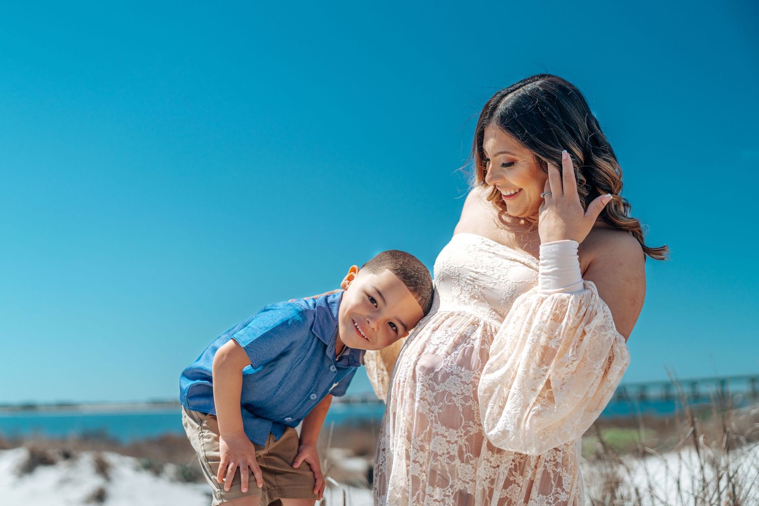 A mother and child share a joyful moment together on a sunny beach with blue skies and sand dunes.