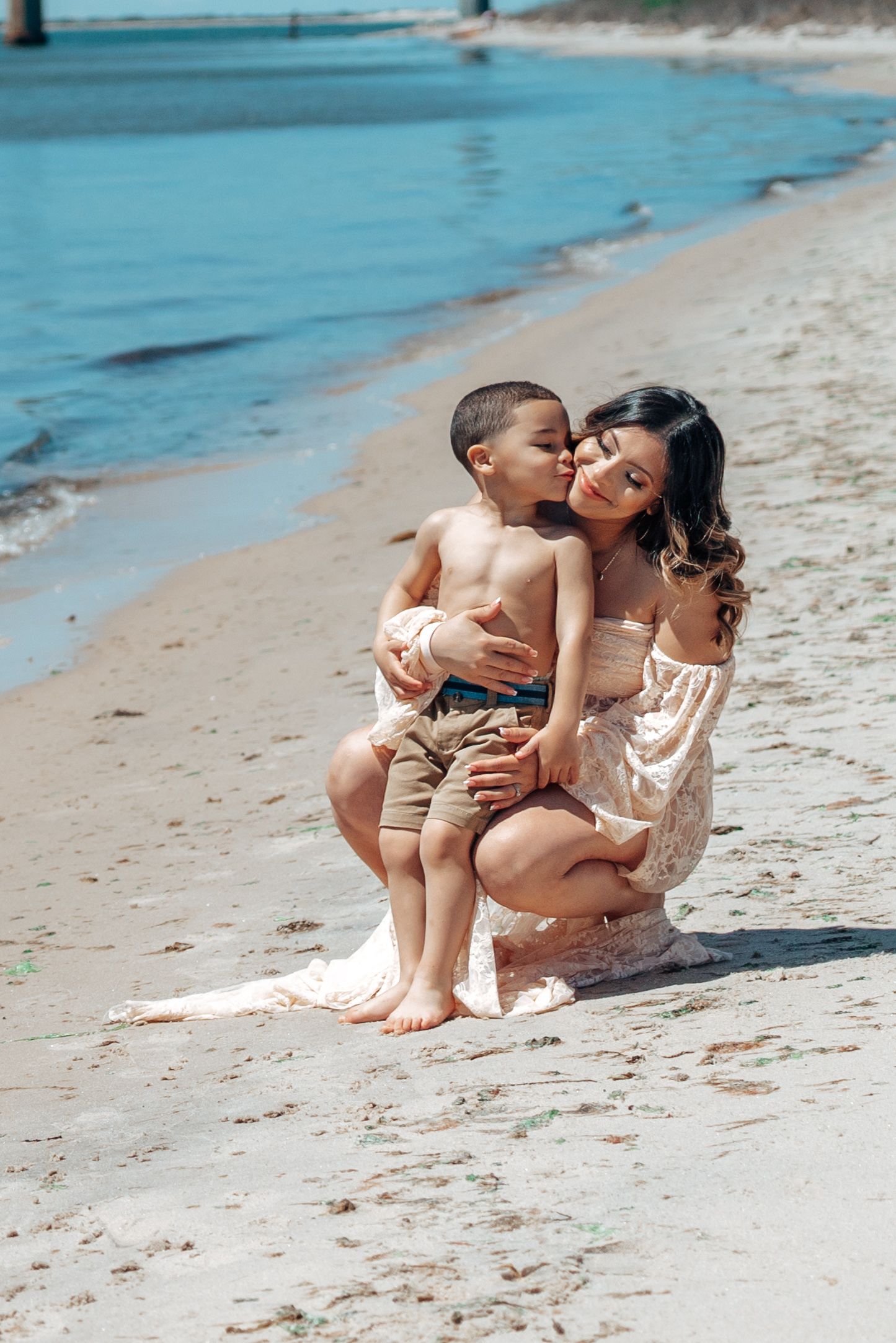 Mother and child share tender moment on sandy beach near ocean waves.