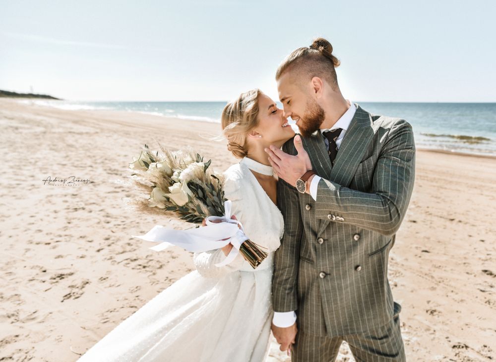 Couple sharing intimate moments on a sunny beach with bridal bouquet.