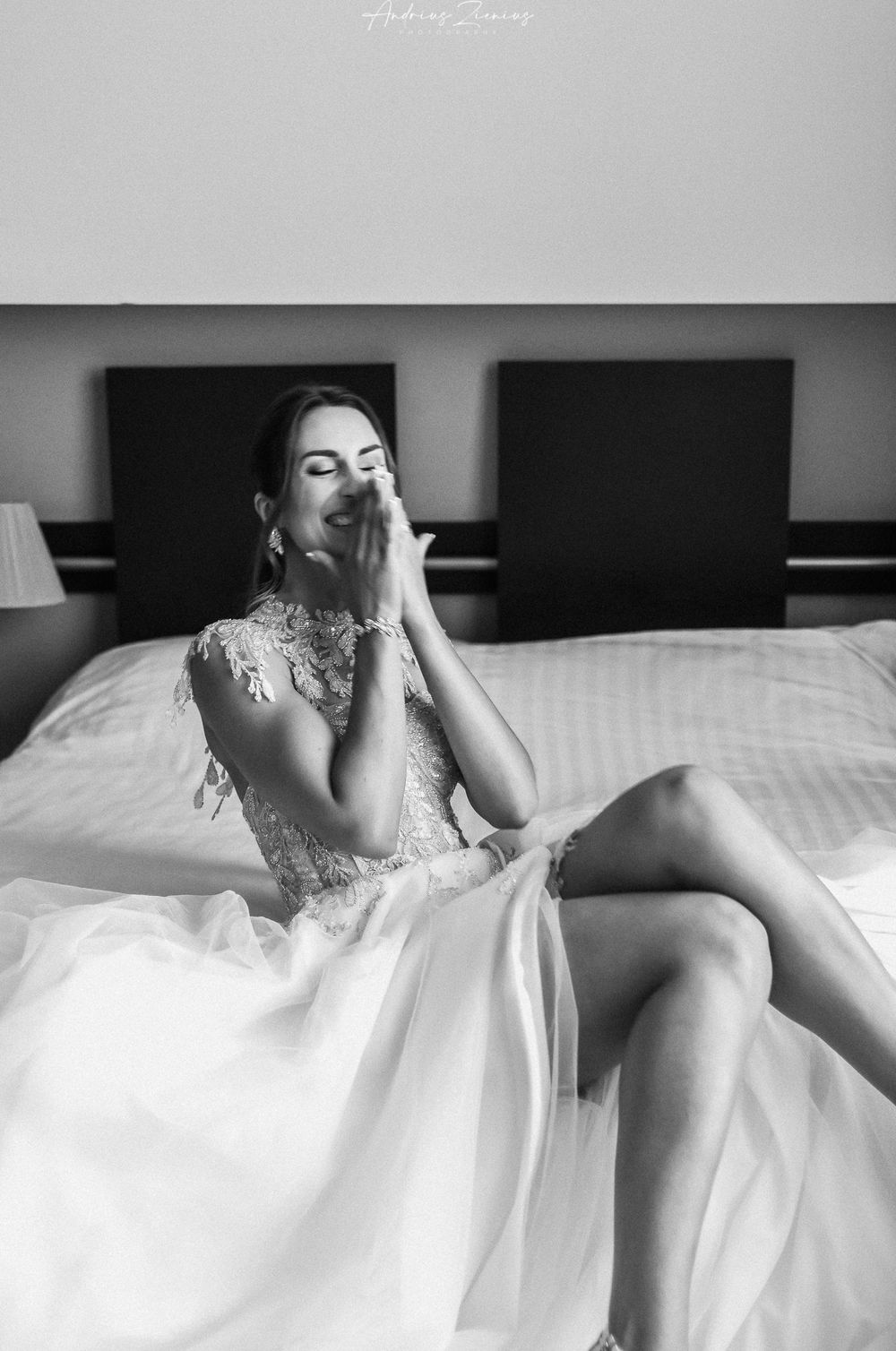A bride in a lace wedding dress sits elegantly on a bed in a black and white portrait.