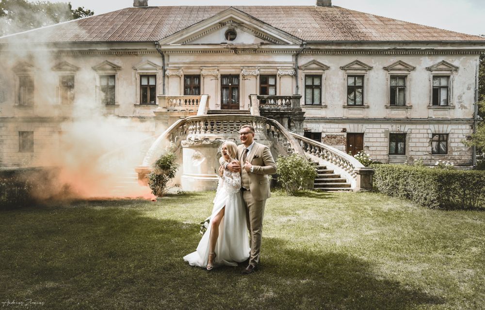 Newlyweds pose romantically in front of a historic mansion with colorful smoke effects in the background.