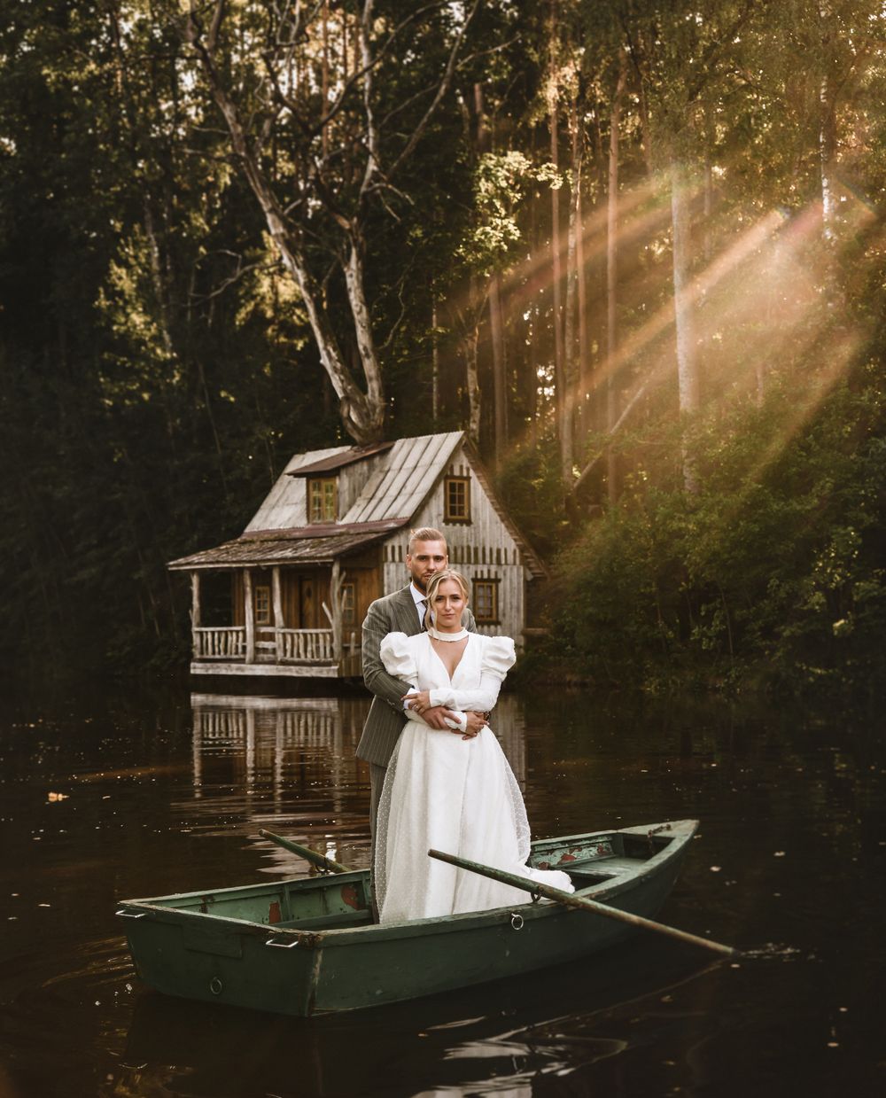 Wedding photo of couple in a rowboat by a charming lakeside cabin with sunbeams through trees.