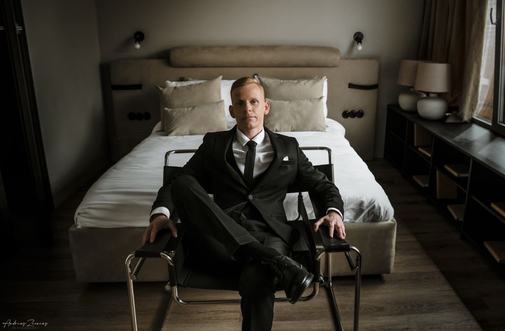 Groom in dark suit seated in modern hotel room before wedding ceremony.