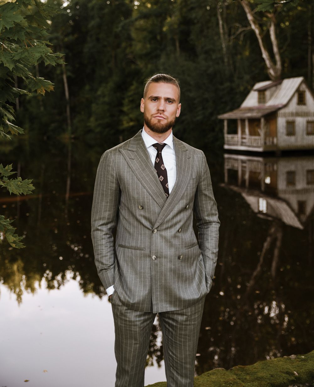 Groom in pinstripe suit poses by reflecting pond with rustic cabin in background.