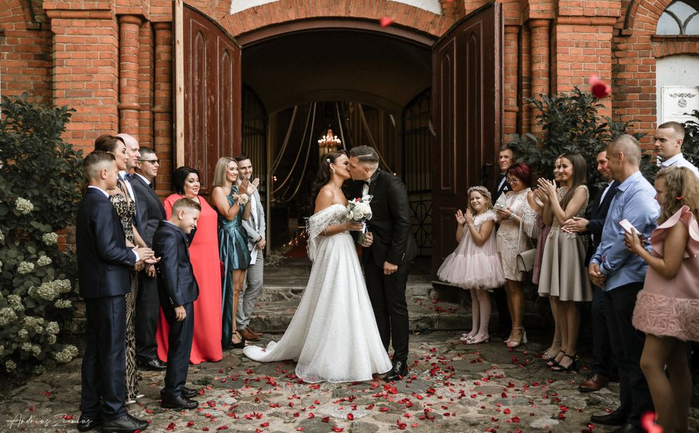 Guests throw rose petals as newlyweds exit a brick church with arched doorways.